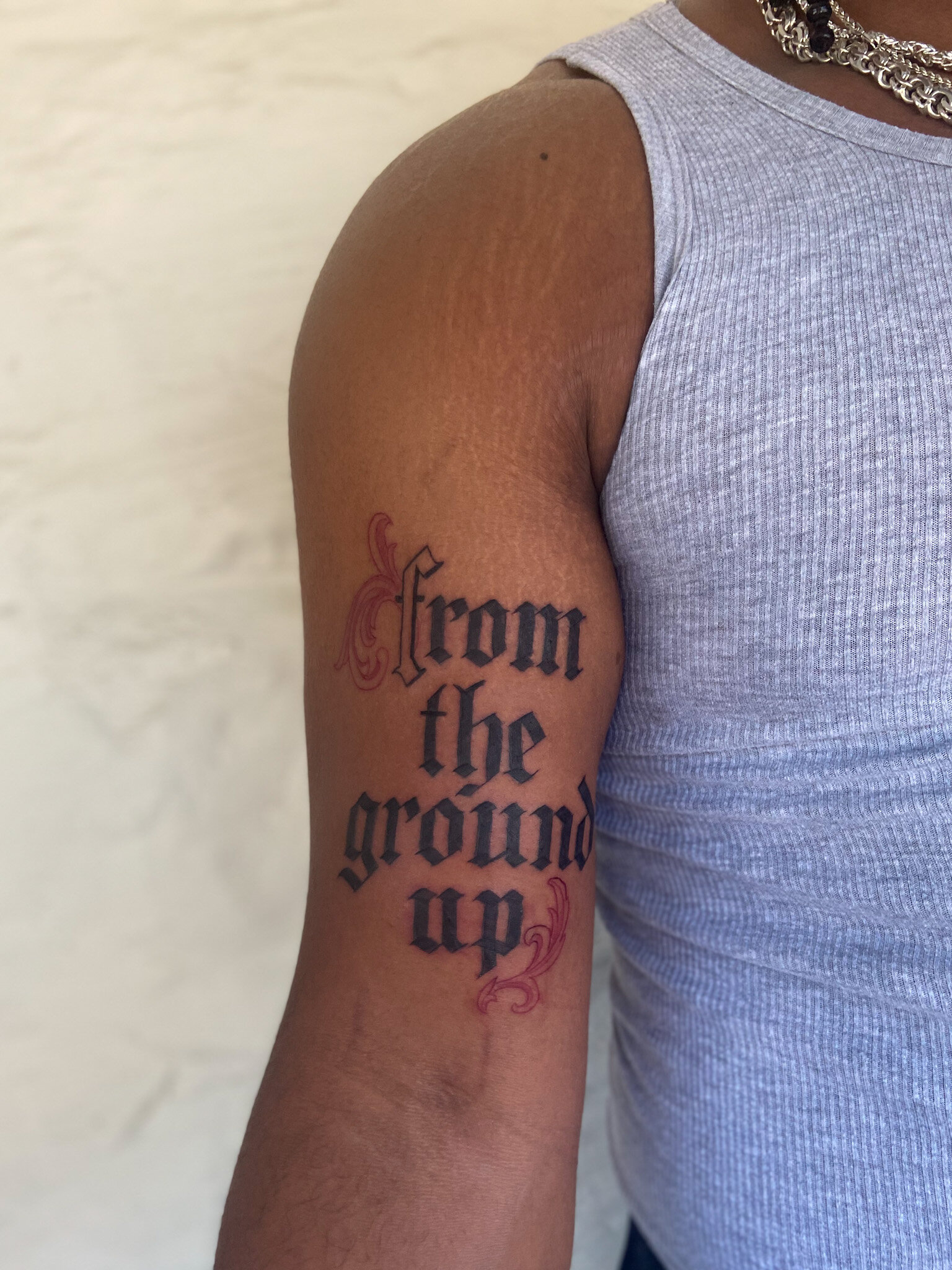 Black letter/ old English style calligraphy tattoo. The lettering tattoo is on a very muscular bicep of a man with deep melanin skin tone. The tattoo says “from the ground up” with a red flourishing design.  