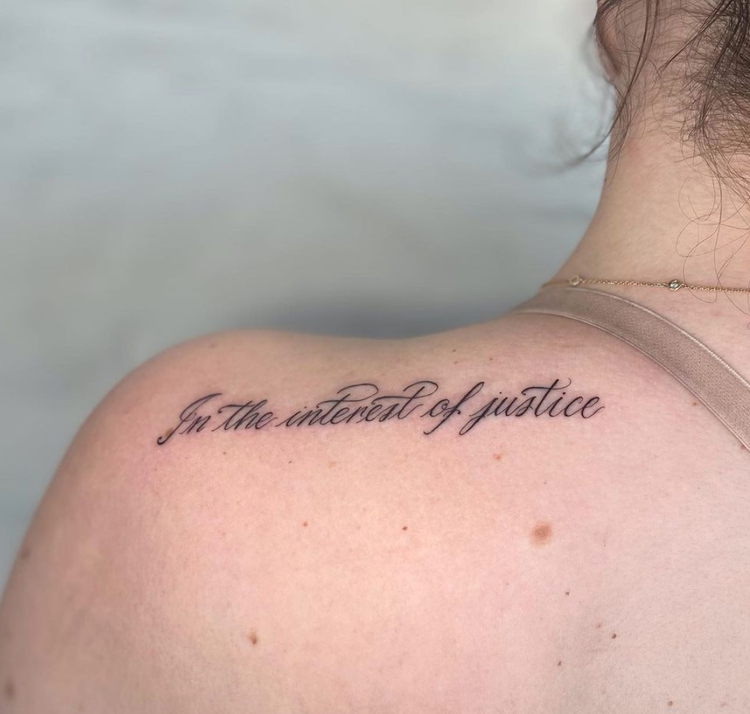  small script tattoo on back of left shoulder. Reads, “In the interest of justice.” 