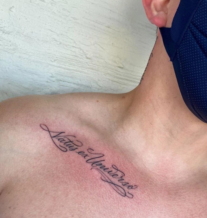  script lettering tattoo along a collar bone. tattoo says “Natus ex Universa” 