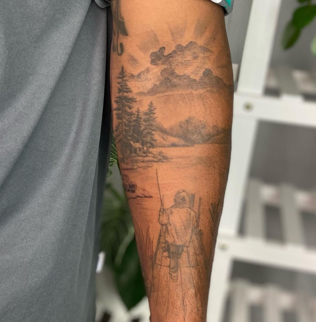  Black and gray, fine line tattoo of a scene including the fisherman’s son on a dock, looking out at the lake, mountains, sky and trees. 