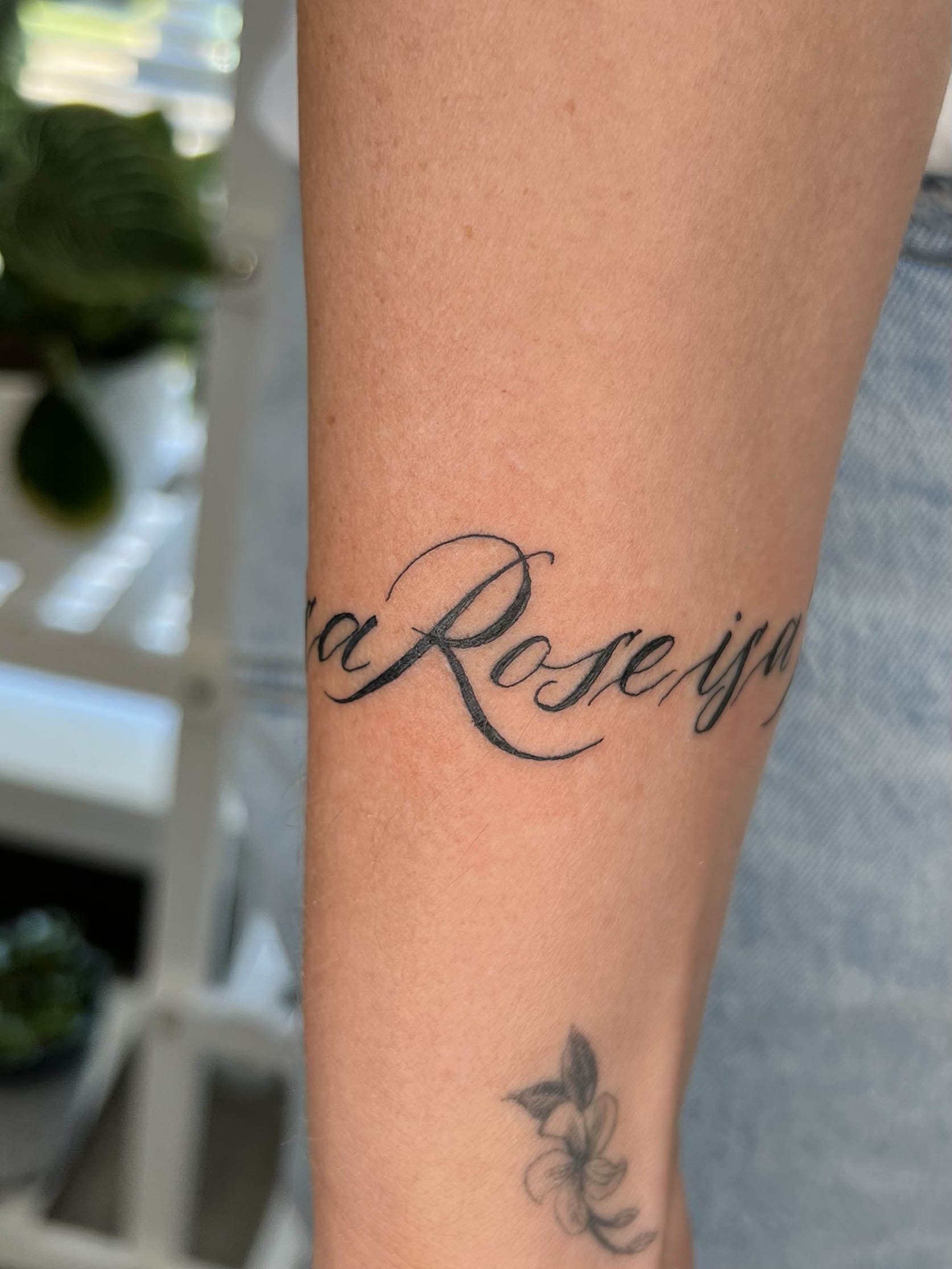  wrap around script around the lower forearm of a woman with neutral toned skin. Calligraphy is drawn by hand and says a Rose is a Rose is a Rose 