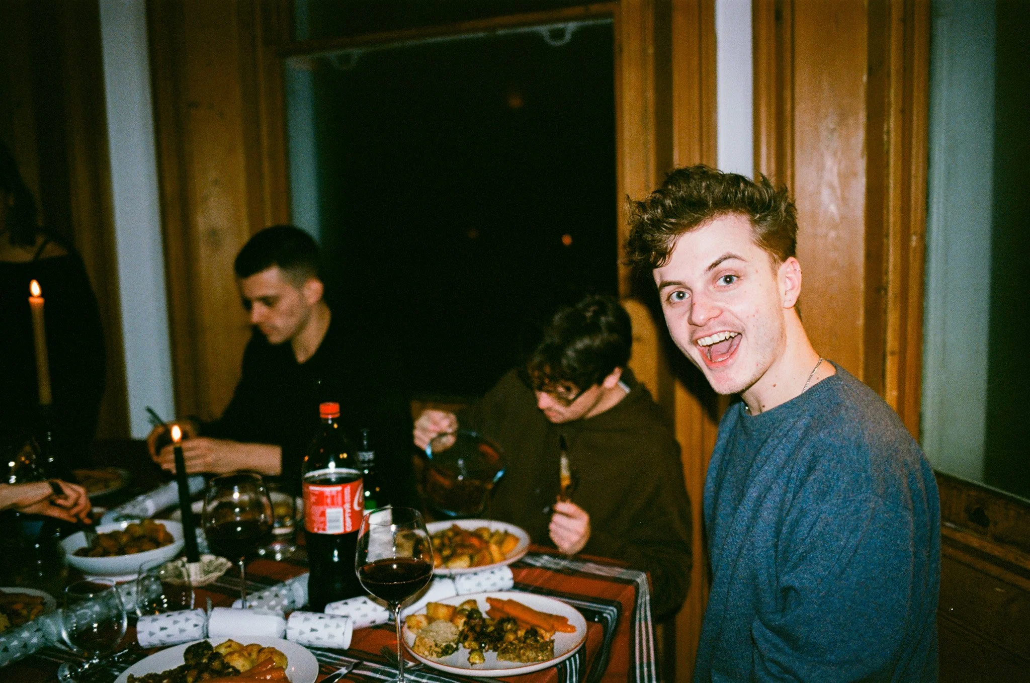 Young man smiling at camera during dinner party with others dining at a table with food and drinks.