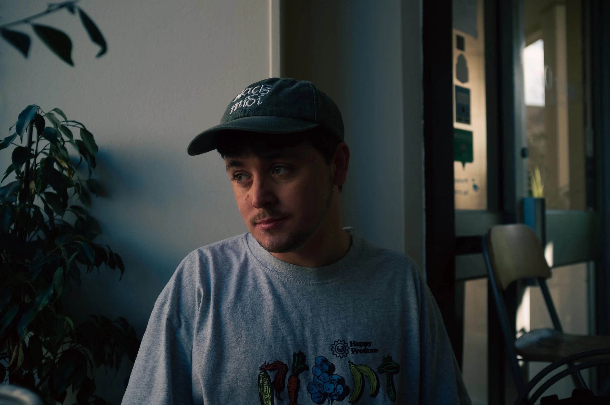 A young man with a light beard and mustache wearing a black baseball cap and a grey t-shirt sitting inside a building near a wall, with a potted plant to his left and sunlight coming through the windows in the background.