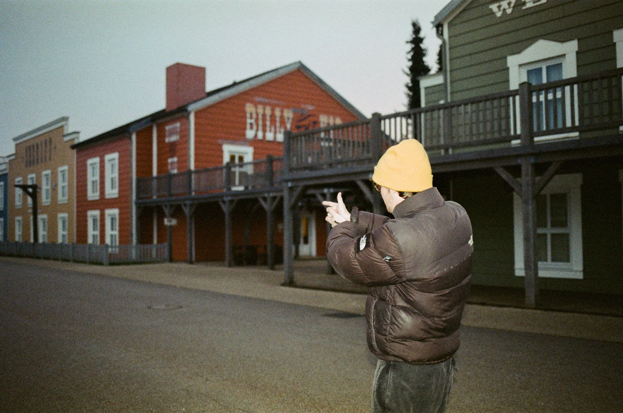 A person wearing a yellow beanie and brown jacket taking a photo with a smartphone on a street lined with colorful buildings.