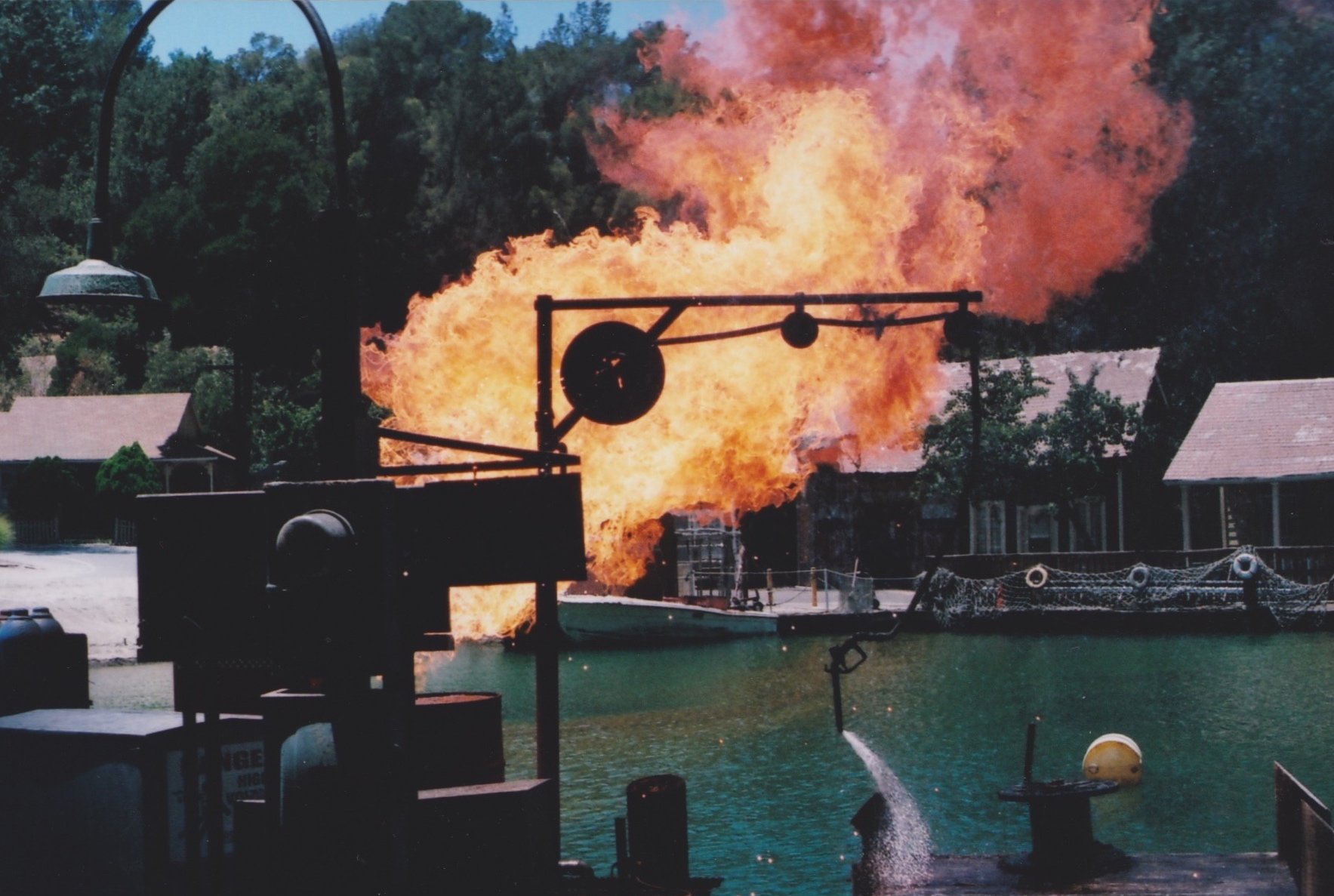 A large explosion or fire on a boat or watercraft, with flames and smoke erupting, near a body of water with trees and buildings in the background.