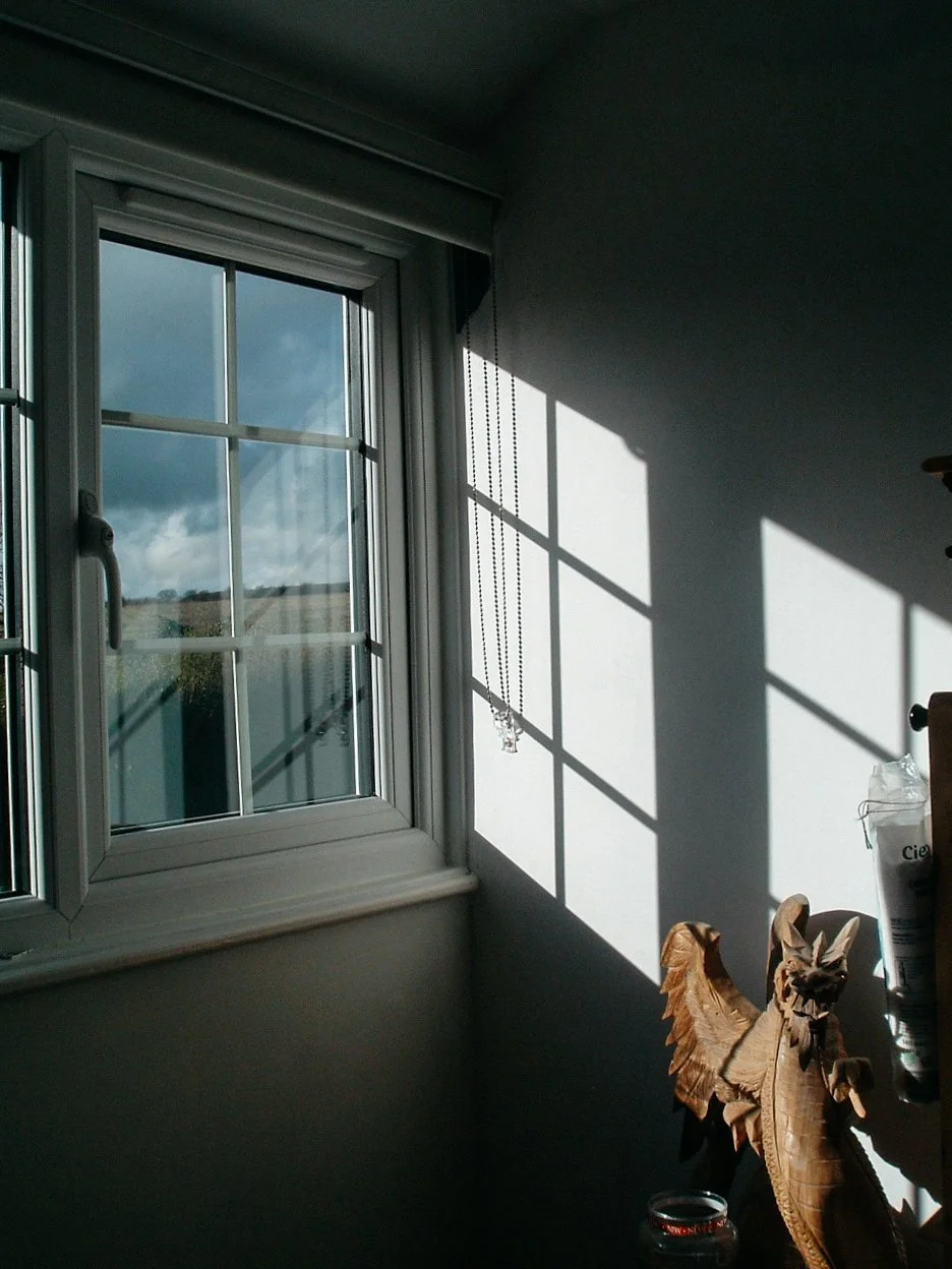 Sunlight casting shadows of a window's frame and blinds onto a wall near a window with an open latch in a room