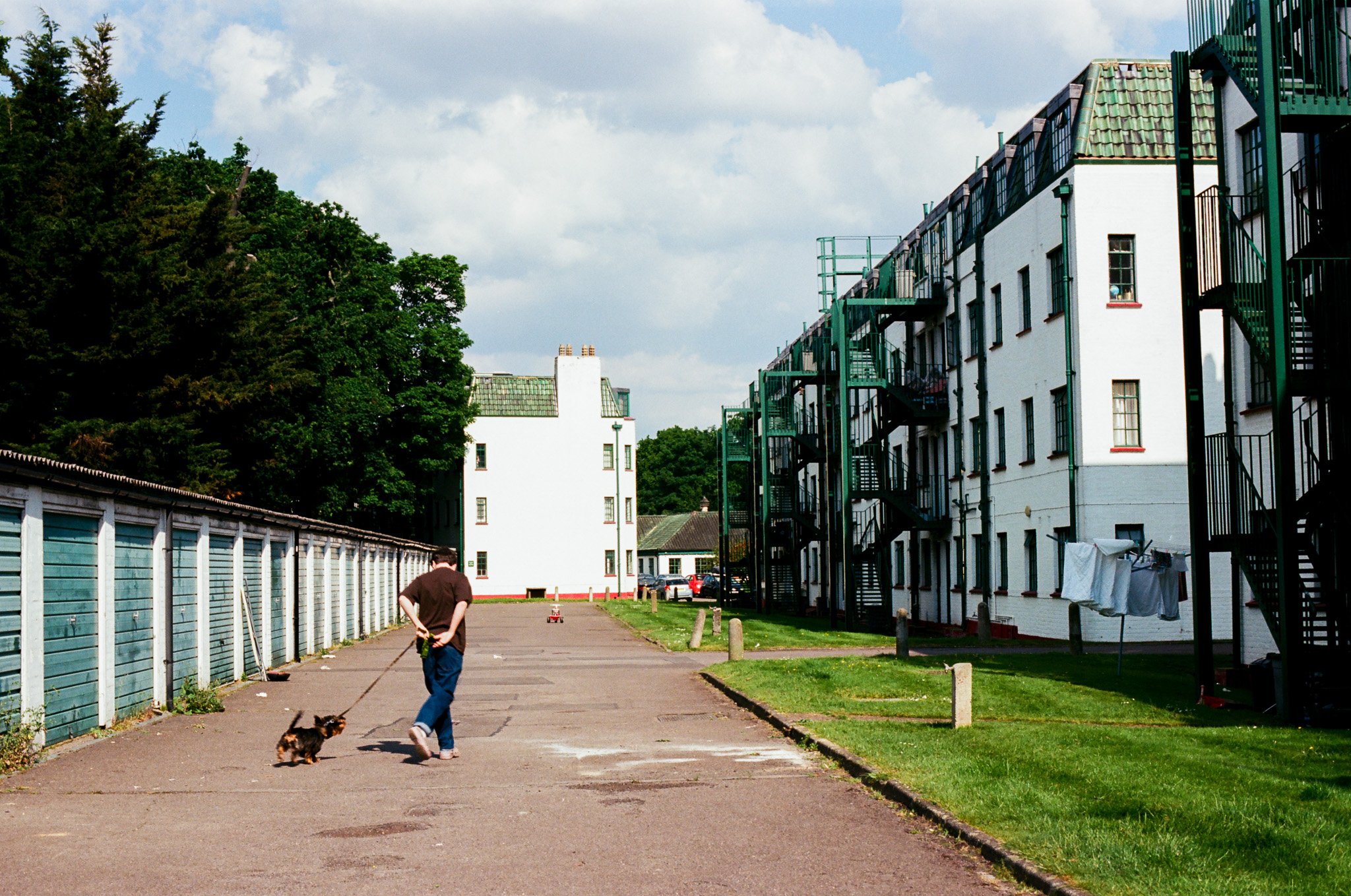 A person walking a small dog along a paved path in front of white apartment buildings with external fire escapes, with trees and a partly cloudy sky in the background.