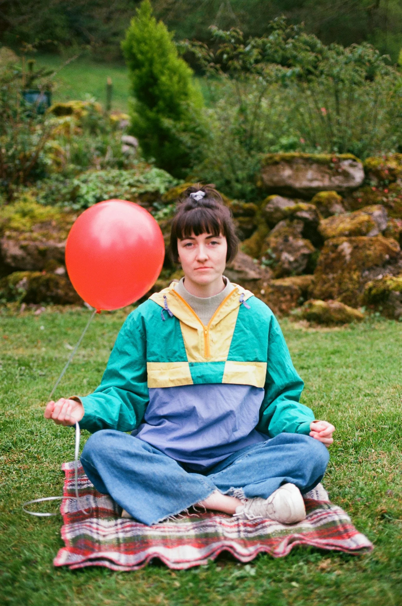 A young woman sitting cross-legged on a plaid blanket outdoors, holding a red balloon in her right hand, with a serious expression, surrounded by lush greenery and rocks.