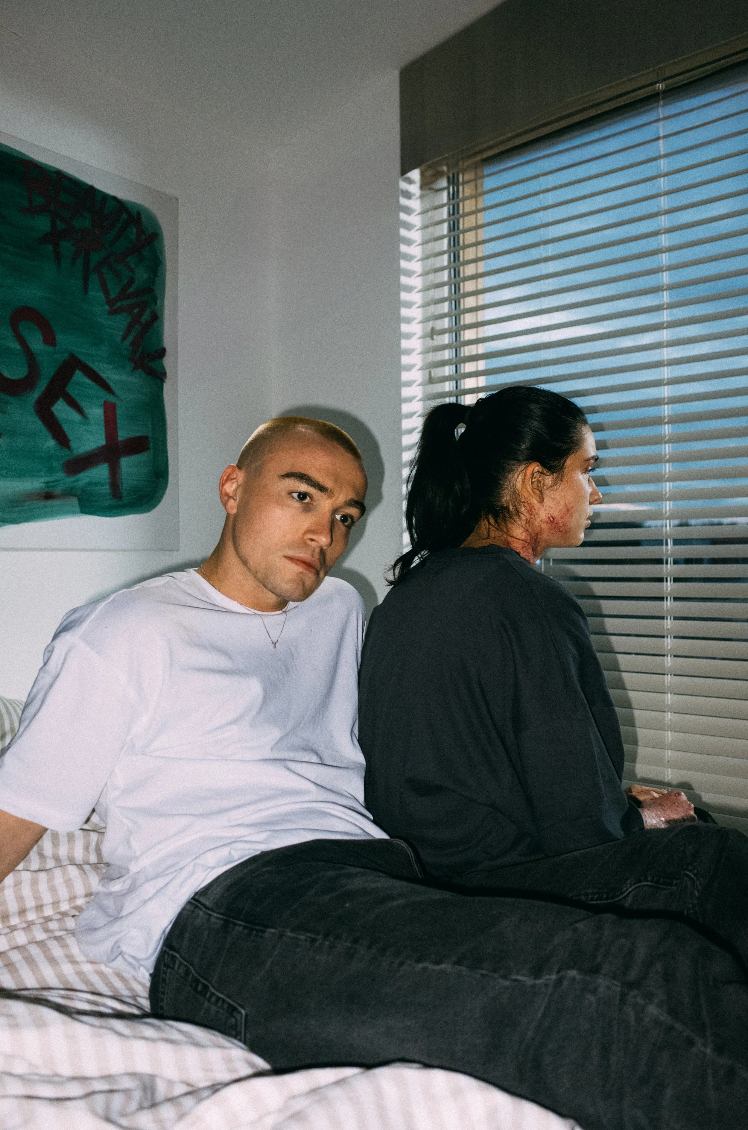A man with a shaved head and a woman with long dark hair, sitting on a bed next to each other, facing away from the camera. The woman has visible facial injuries or scars on her neck and face, and they are looking out a window with blinds. There is a