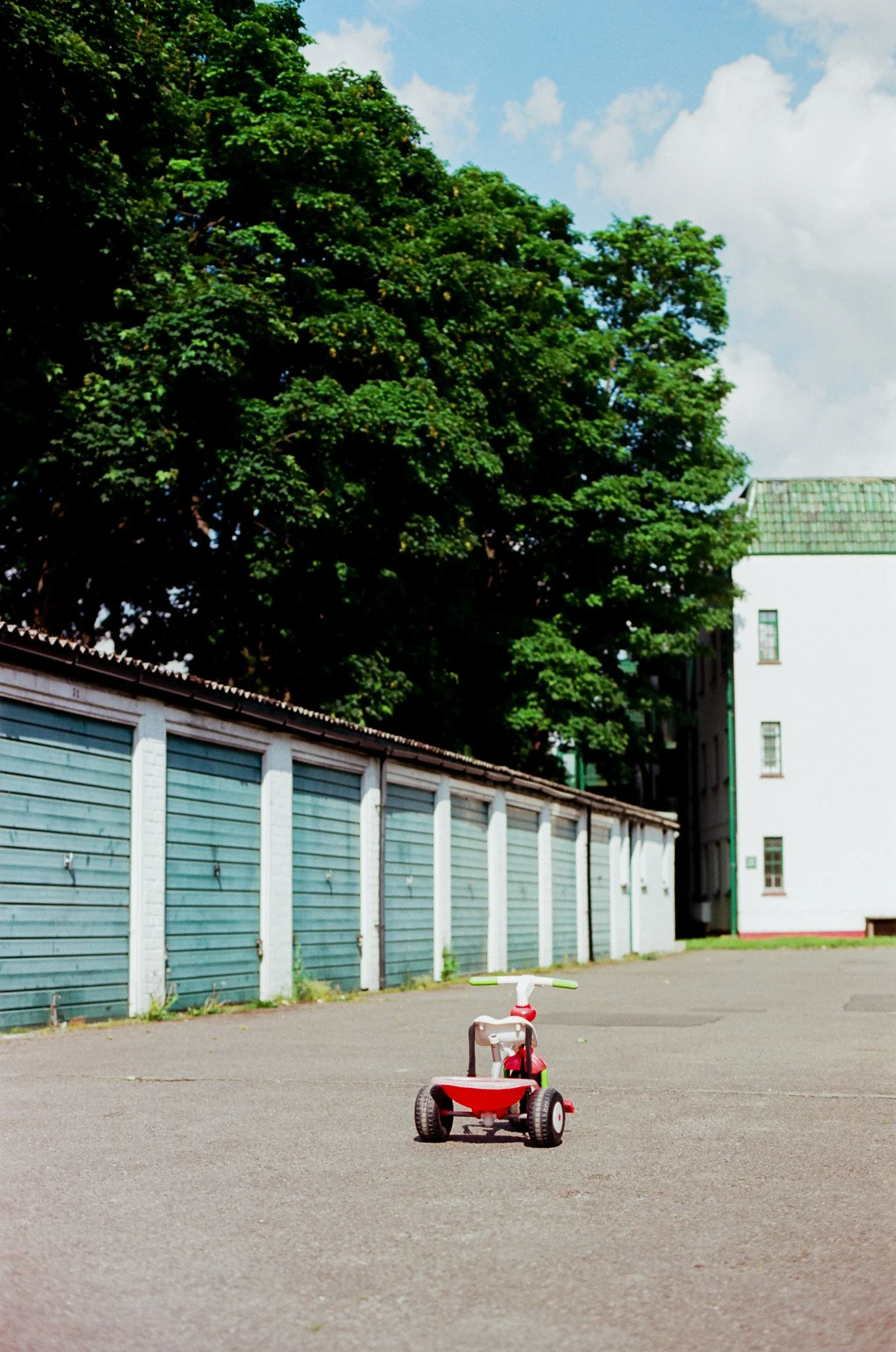 A small red and white children's tricycle parked on an empty paved area, with a large green tree and a building with a white wall in the background under a blue sky with some clouds.