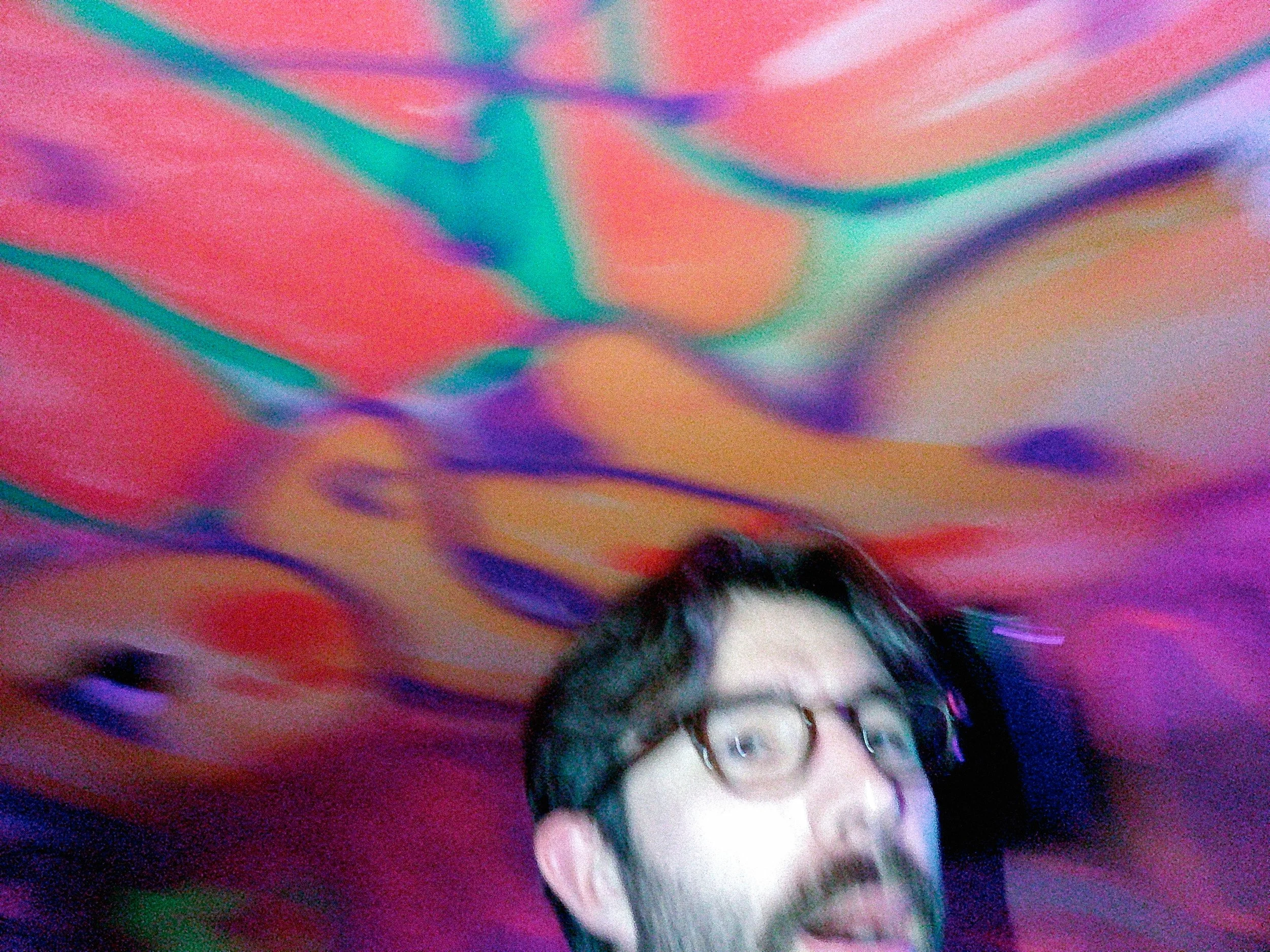 A person with glasses and facial hair is holding their head and appears to be surprised or excited, with a colorful, swirling abstract background.