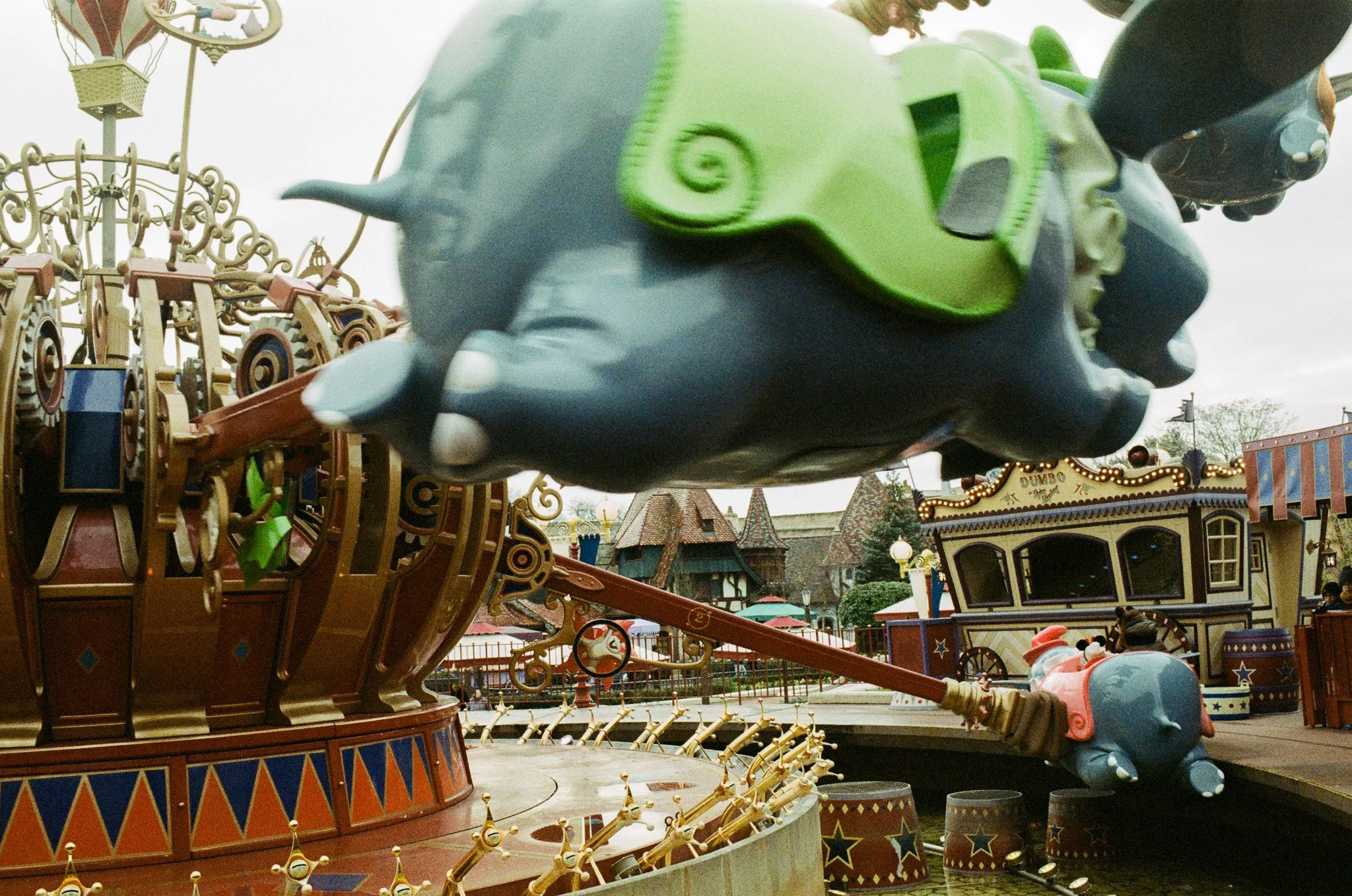 Close-up of a Dumbo the Flying Elephant ride at Disneyland, showing a large elephant figure with green goggles and a pink hat, part of a carousel with small elephant figures attached to a red and gold structure, surrounded by other amusement park att