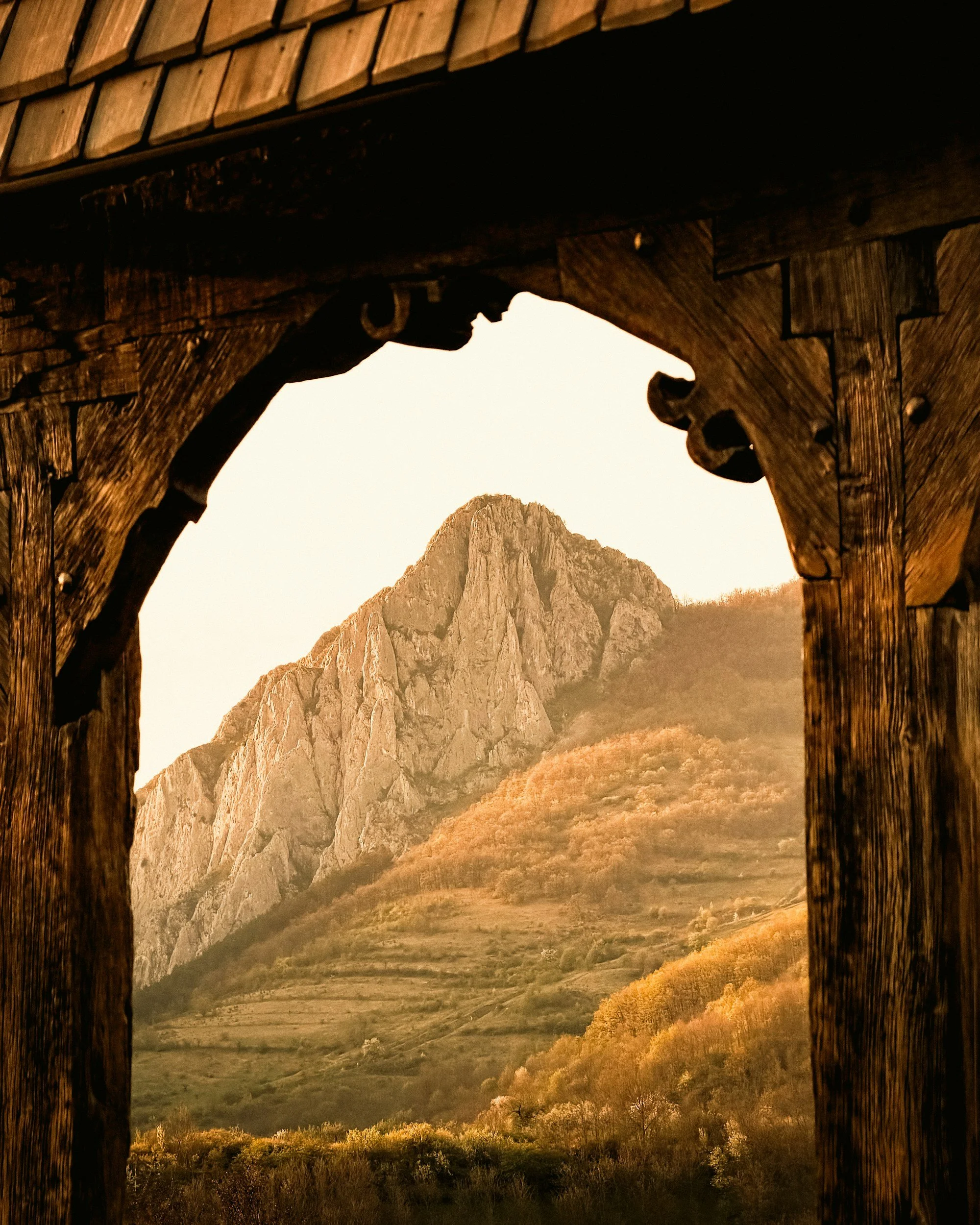 Wooden structure framing a mountain landscape with large rocky peaks and colorful hillsides during sunset.