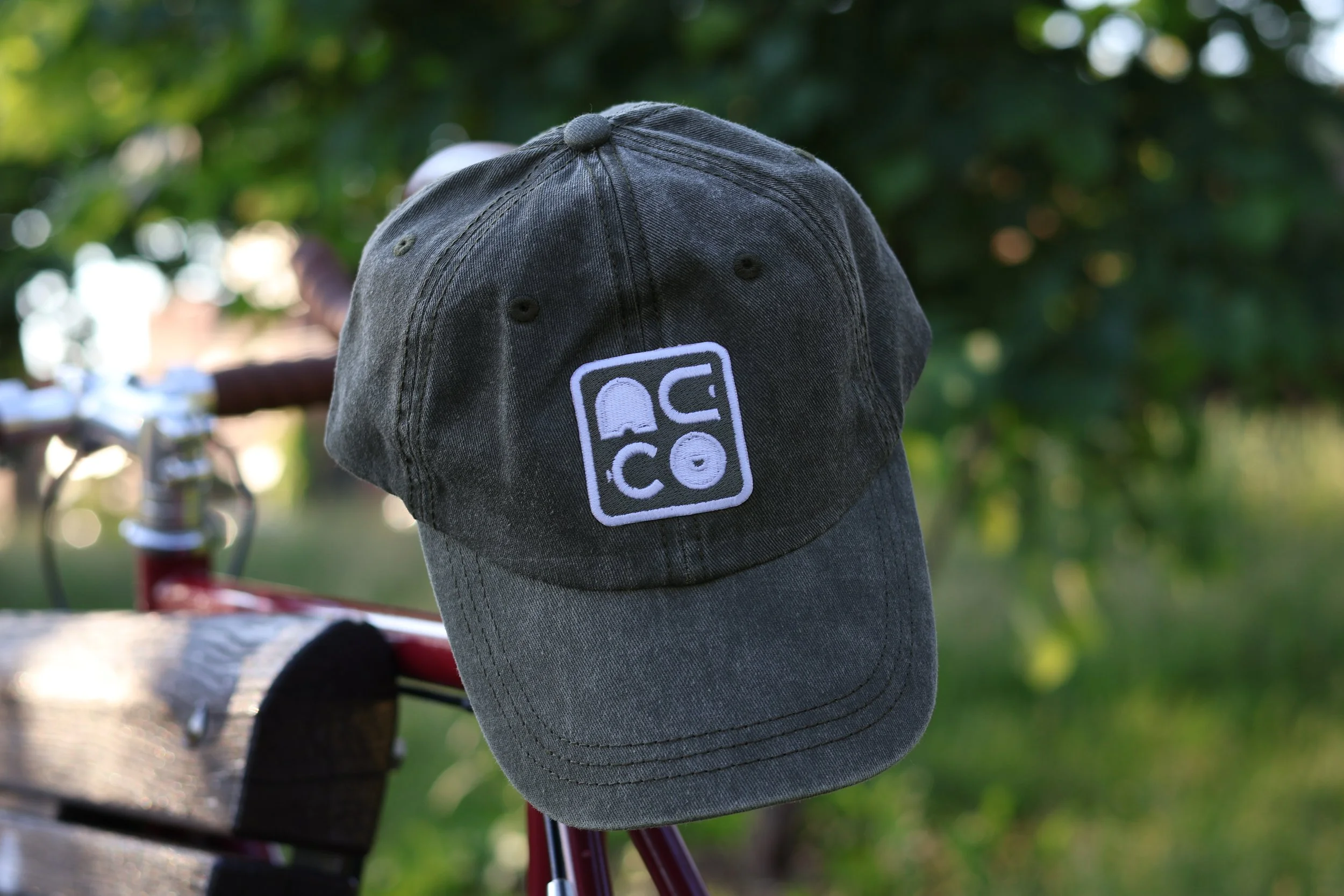 Un casquette verte avec le logo Neko brodé posée sur le guidon d'un vélo rouge, dans un parc avec des arbres en arrière-plan.