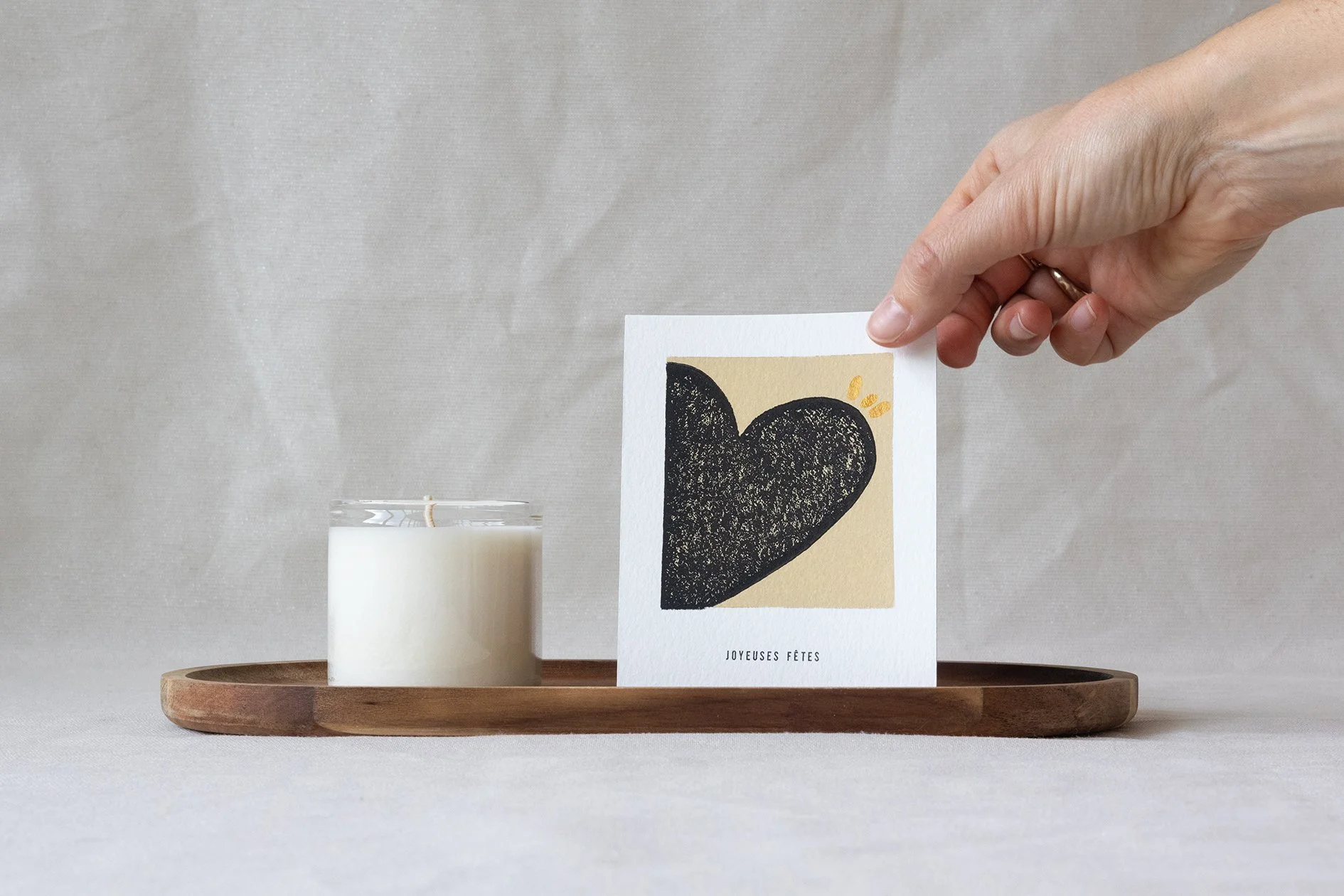 Une main tient une carte de vœux avec un dessin d'un cœur noir imprimé avec la technique de la linogravure. La carte est posée au côté d'une bougie sur un plateau en bois avec un arrière plan beige.