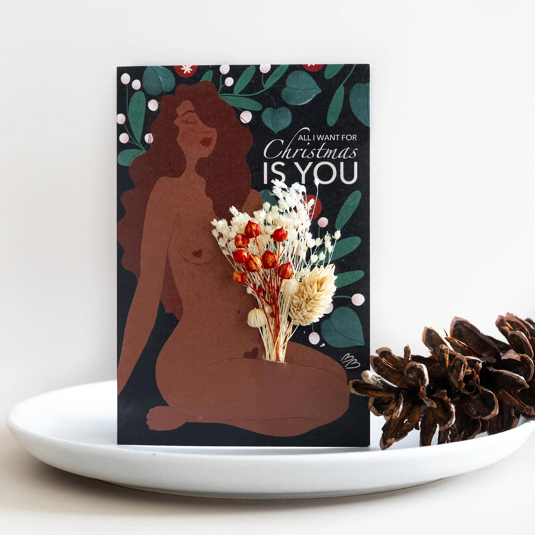 Carte de Noël avec une illustration d'une femme nue et un bouquet de fleurs séchées en première plan, accompagnée de pommes de pin sur une assiette blanche.