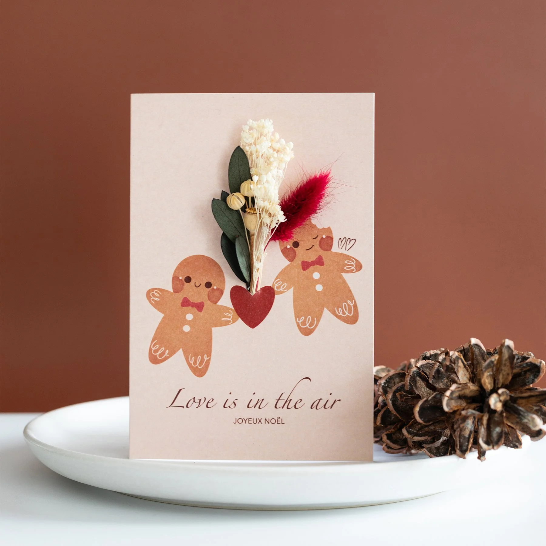 Carte de vœux de Noël avec deux bonhommes en pain d'épice, un cœur rouge et des fleurs séchées. La carte est disposée sur une assiette blanche agrémentée d'une pomme de pin.