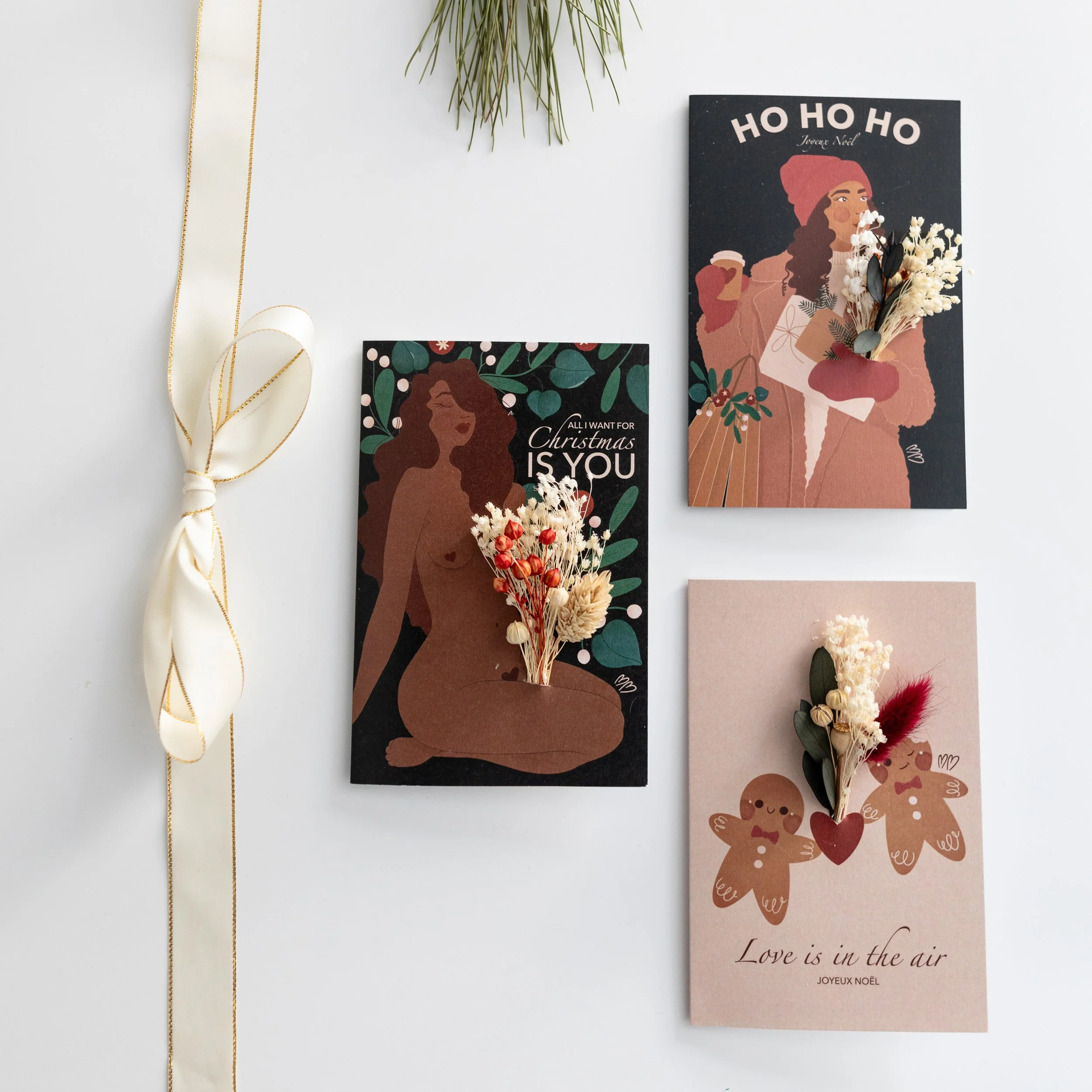 Collection de trois cartes de vœux de Noël avec des illustrations de femmes ou de bonhommes en pain d'épice, décorées de petites fleurs séchées et de feuilles, disposées sur une surface blanche.