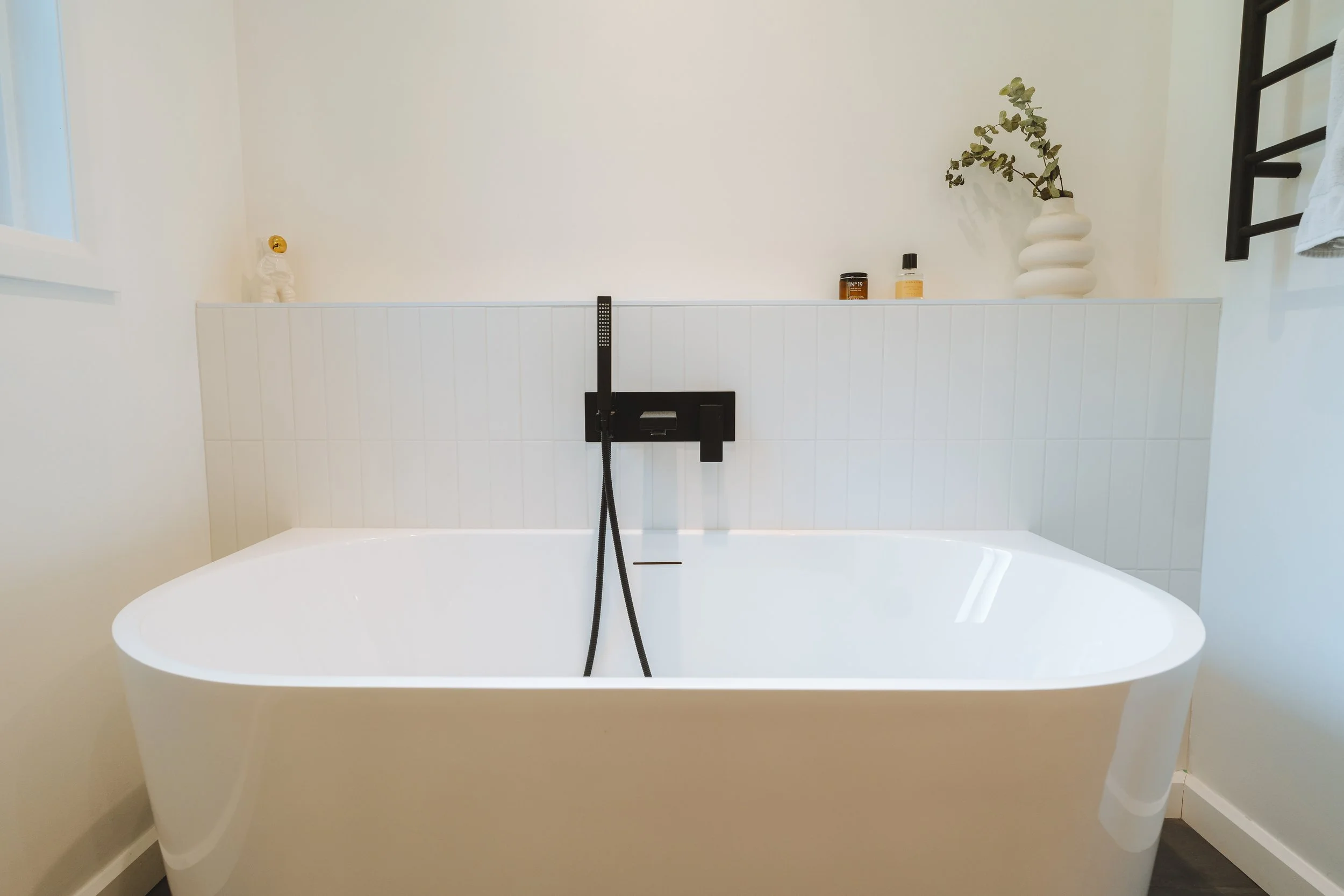 Baignoire blanche dans une salle de bain minimaliste avec quelques objets décoratifs et produits de bain sur une tablette au mur.