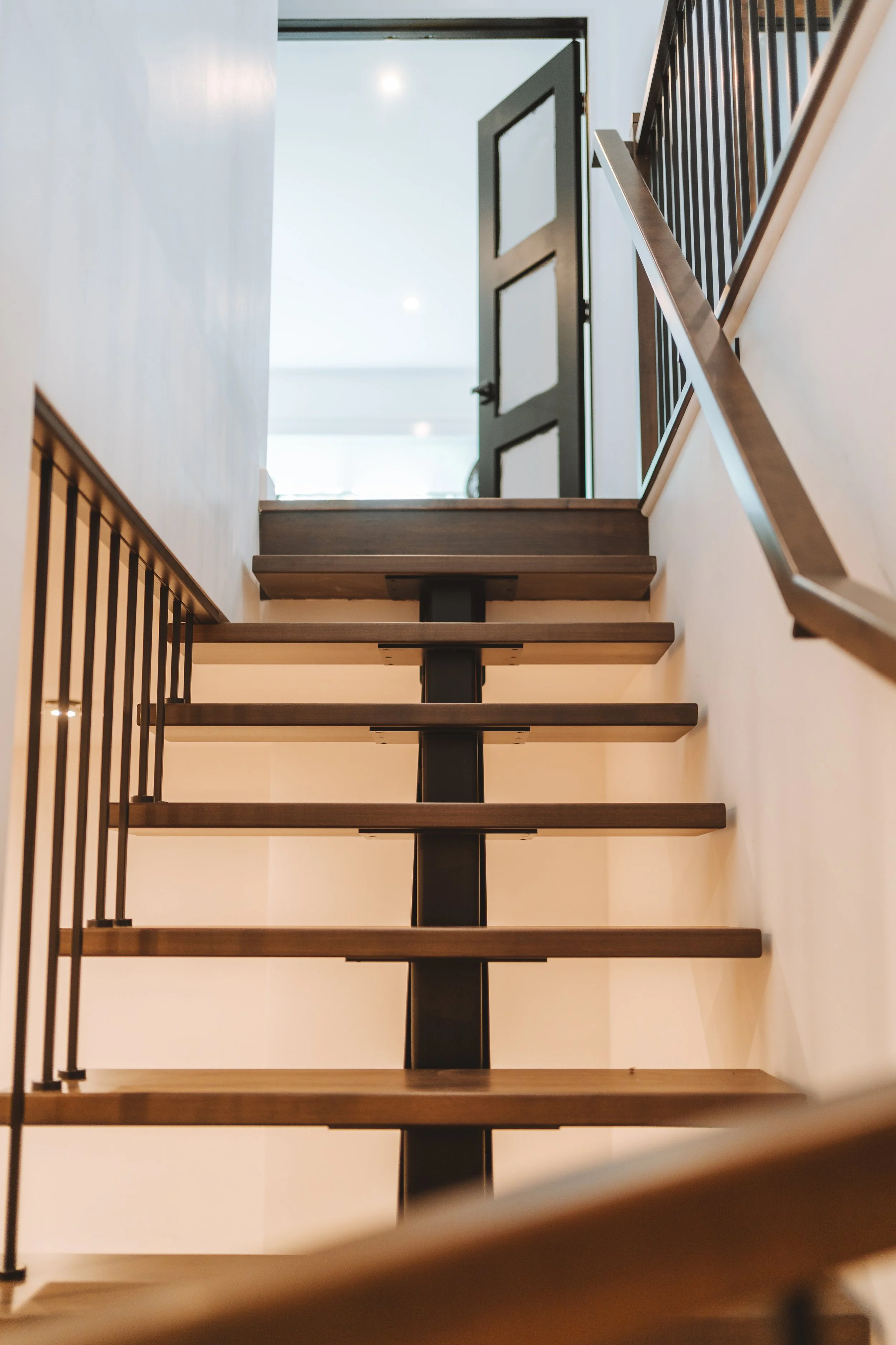 Escalier intérieur en bois avec rampe en métal, vue de bas en montant vers une porte ouverte au plafond lumineux.