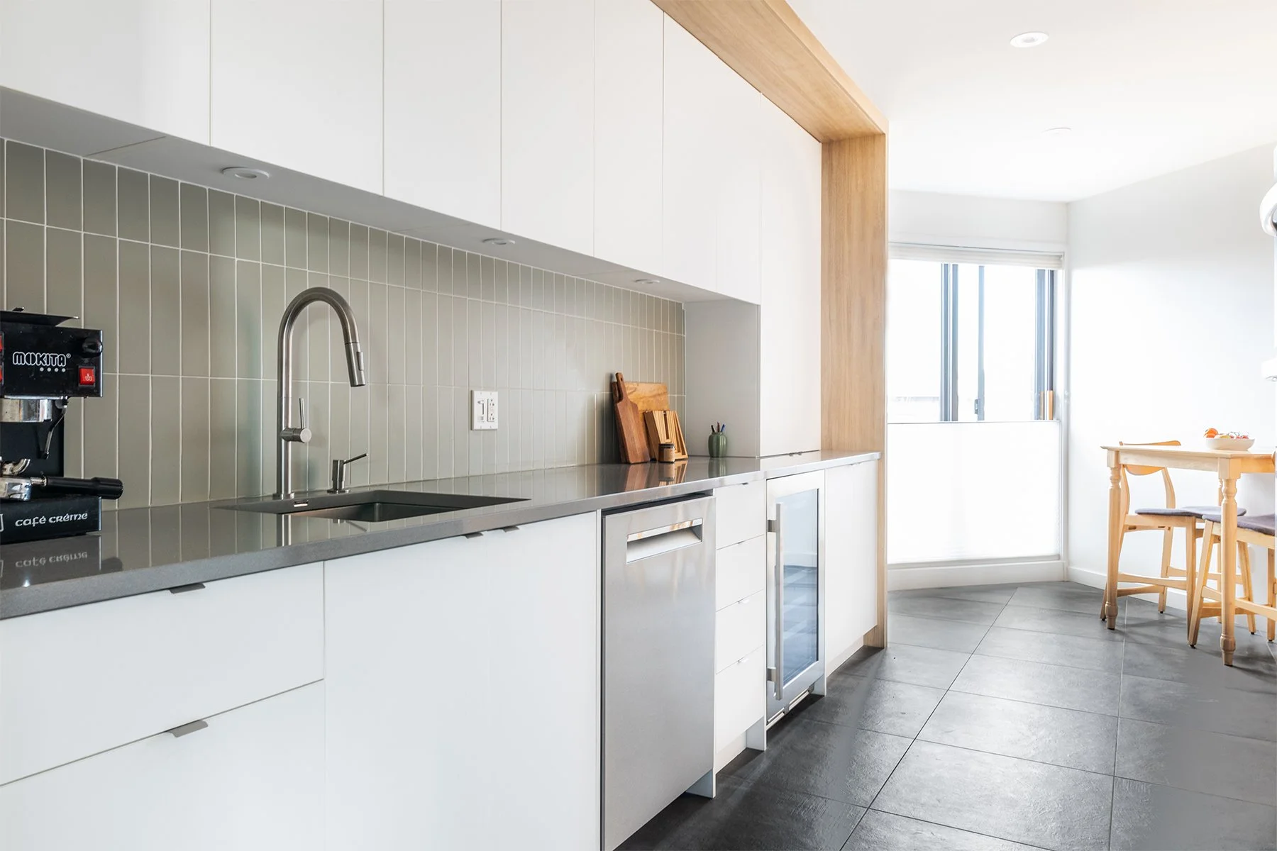 Cuisine moderne avec plan de travail gris, évier en inox, étagère en bois, et mobilier blanc, éclairée naturellement par une fenêtre.