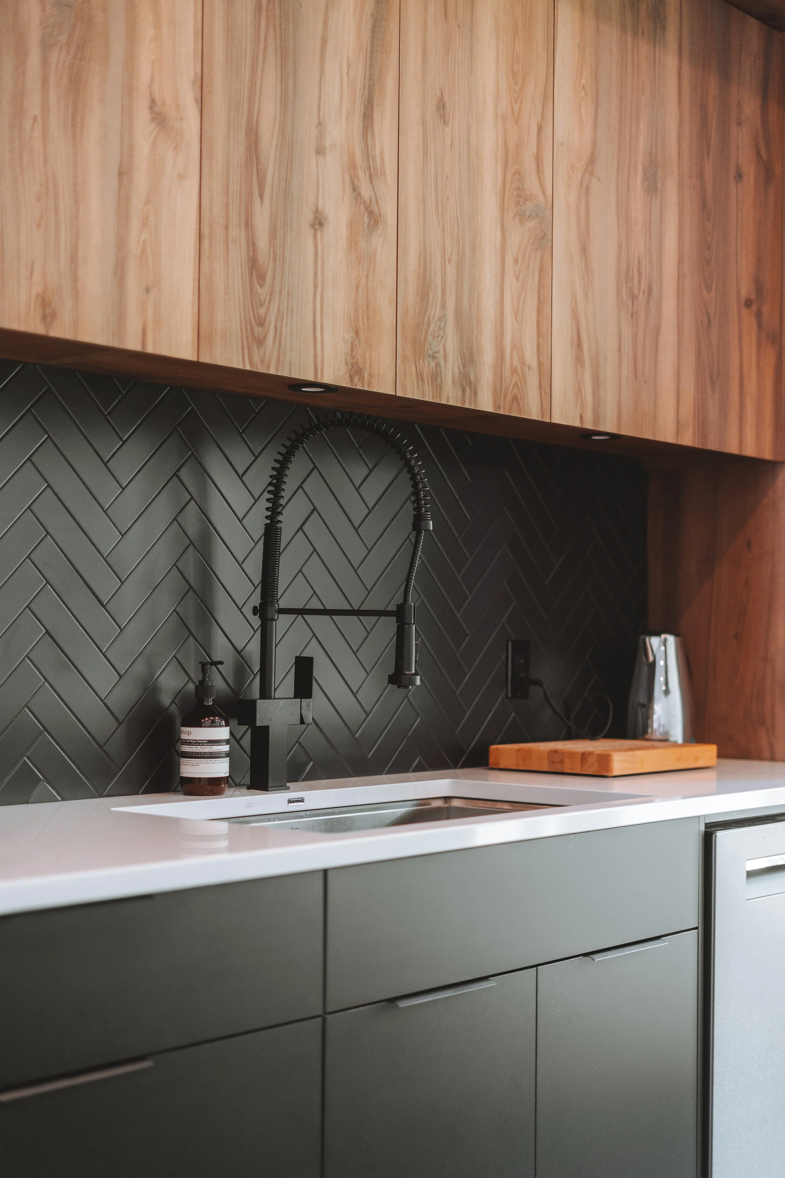 Cuisine moderne avec plan de travail blanc, armoire noire et bois, évier noir avec robinet haut, dosseret en carreaux de métro noirs, accessoires en bois et appareils électroménagers.