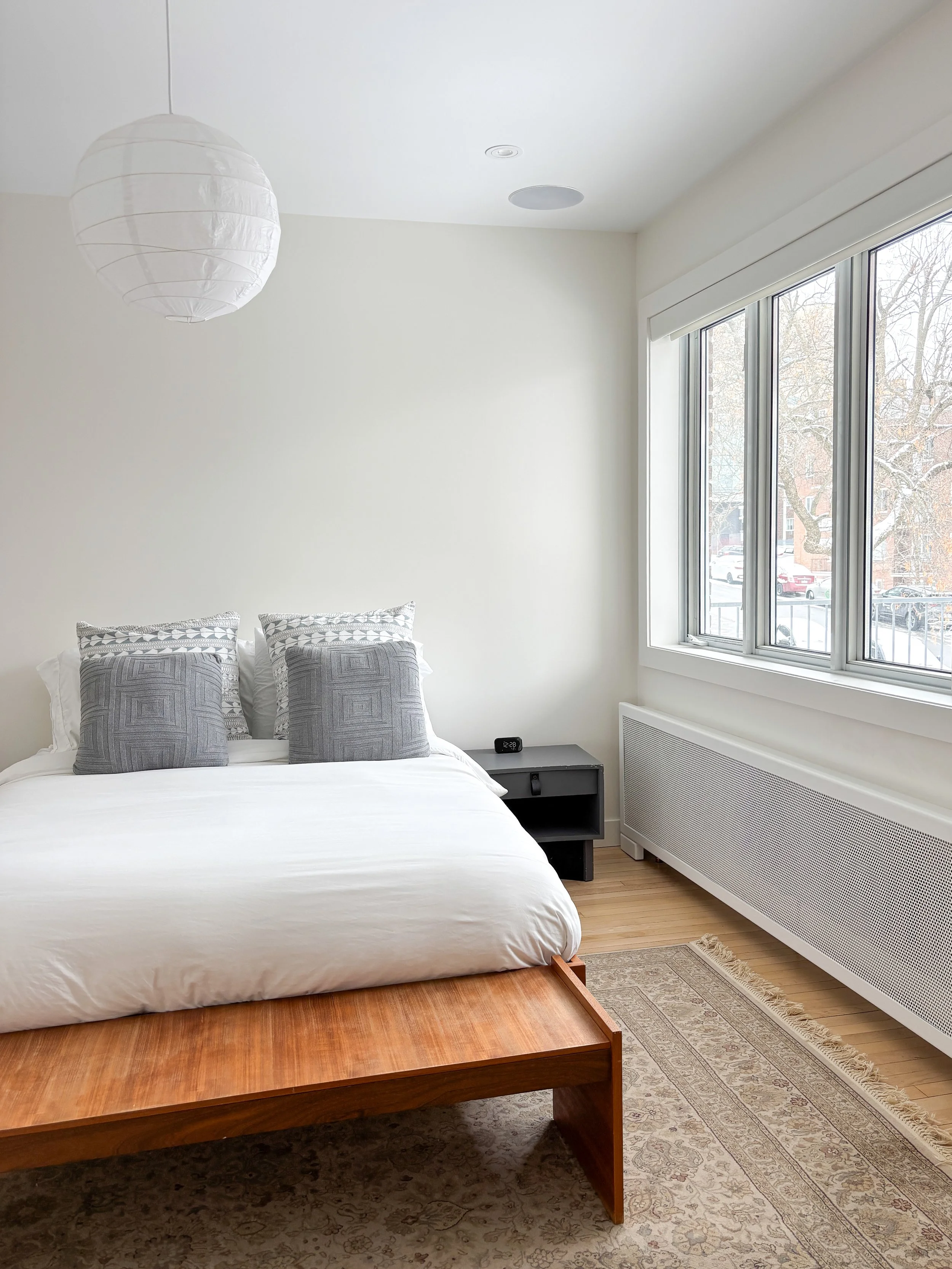 Chambre minimaliste avec lit blanc, quatre coussins gris, petit noire de nuit, grande fenêtre laissant entrer la lumière naturelle, et tapis beige.