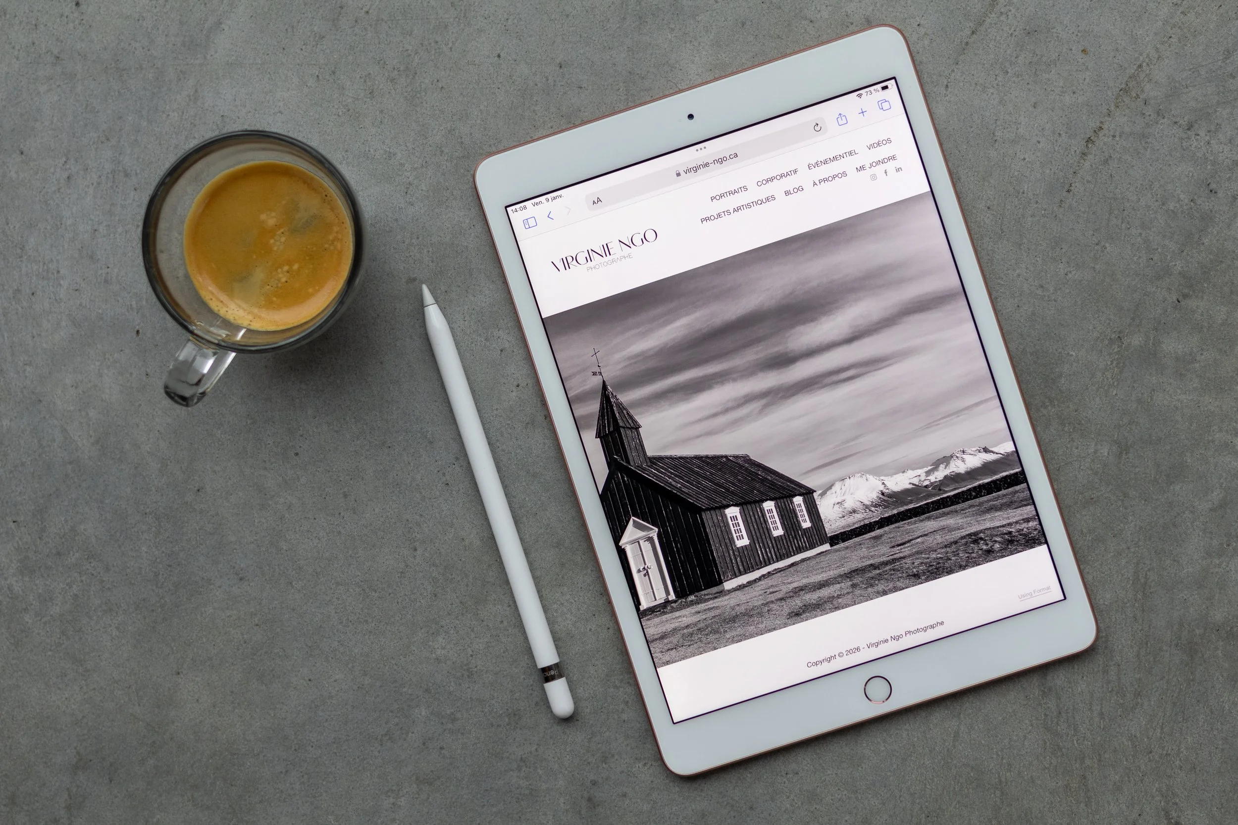 Un iPad affichant le site web de la photographe Virginie Ngo avec une image noir et blanc d'une église en bois dans un paysage montagneux. À côté, un stylo blanc et une tasse de café sur une surface en béton.