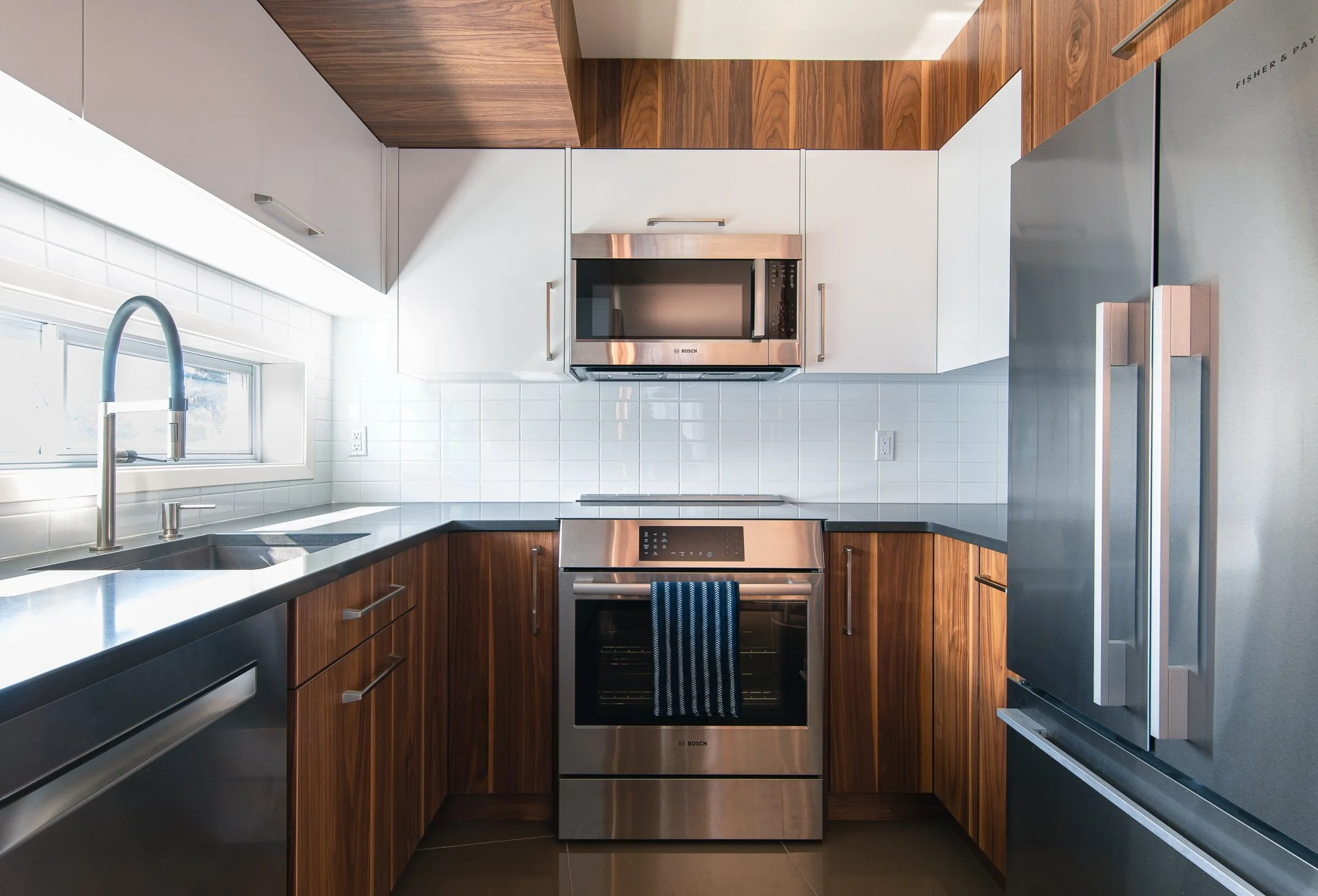 Cuisine moderne avec plan de travail en quartz noir, armoires en bois, carrelage blanc, fenêtre au-dessus de l'évier, four et micro-ondes en acier inoxydable, réfrigérateur en acier inoxydable.