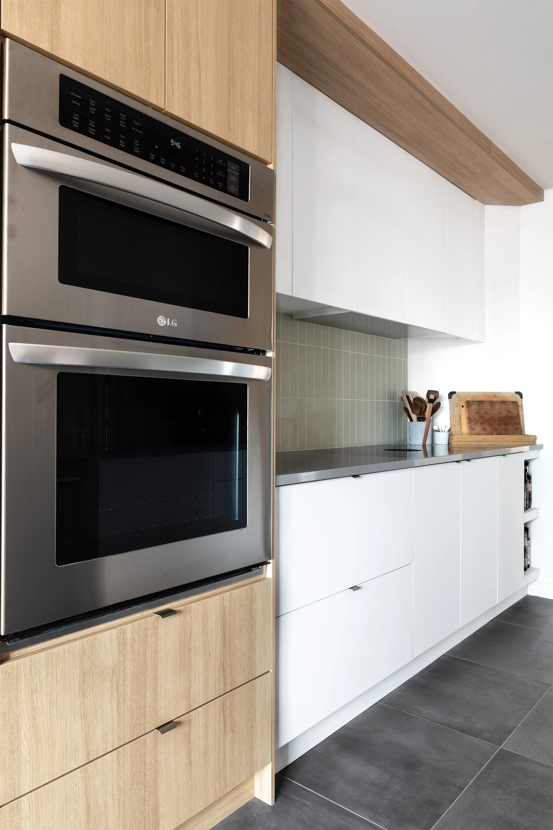 Côté d'une cuisine moderne avec un four et un micro-ondes en acier inoxydable, armoires en bois clair, plans de travail en surface grise, contenant des ustensiles et une planche à découper en bois.