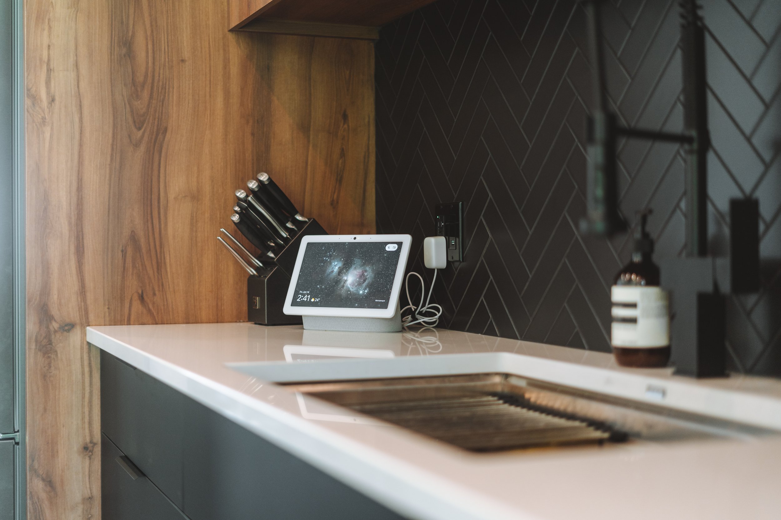 Cuisine moderne avec un plan de travail en bois, une tablette Alexa avec l'écran affichant une galaxie, un bloc de couteaux, un porte-ustensiles blanc, une bouteille d'huile ou de vinaigre et un dispositif noir sur le mur en arrière-plan.