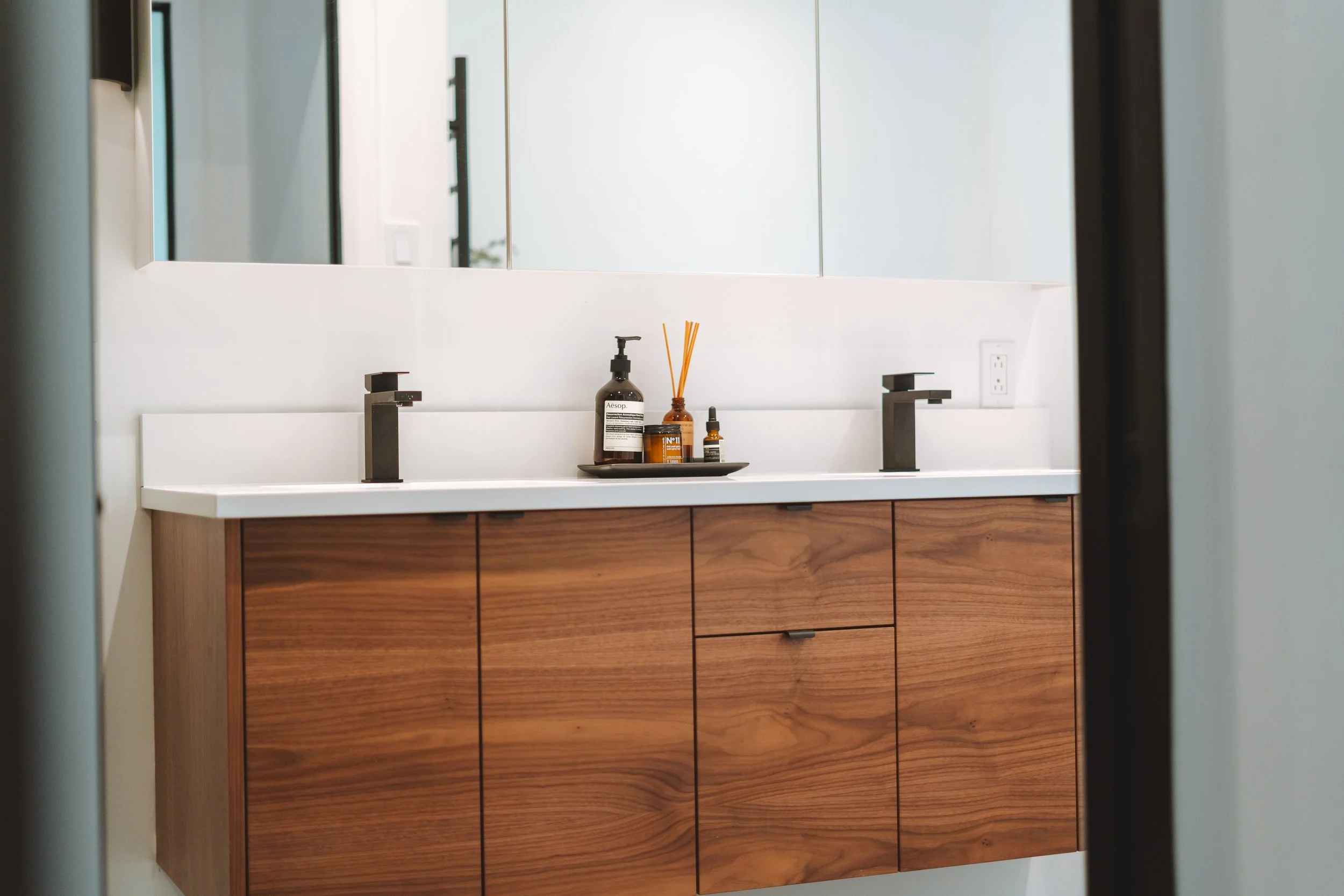 Meuble de salle de bain en bois avec un lavabo double, un grand miroir au-dessus et des bouteilles de produits de toilette au centre.