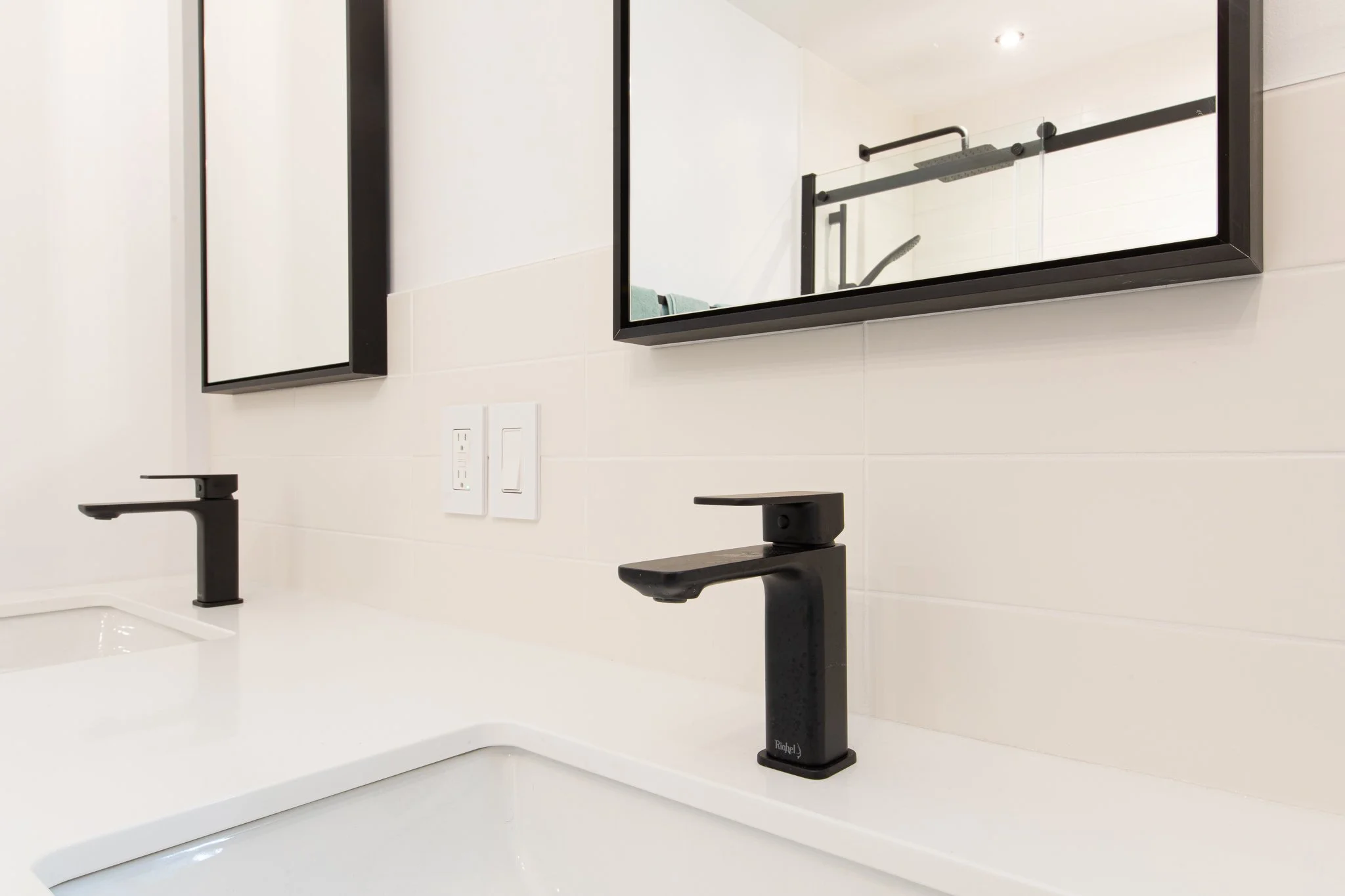 Deux lavabos blancs avec robinets noirs sur un comptoir blanc dans une salle de bain moderne, avec un miroir rectangulaire au-dessus et des prises électriques à côté.