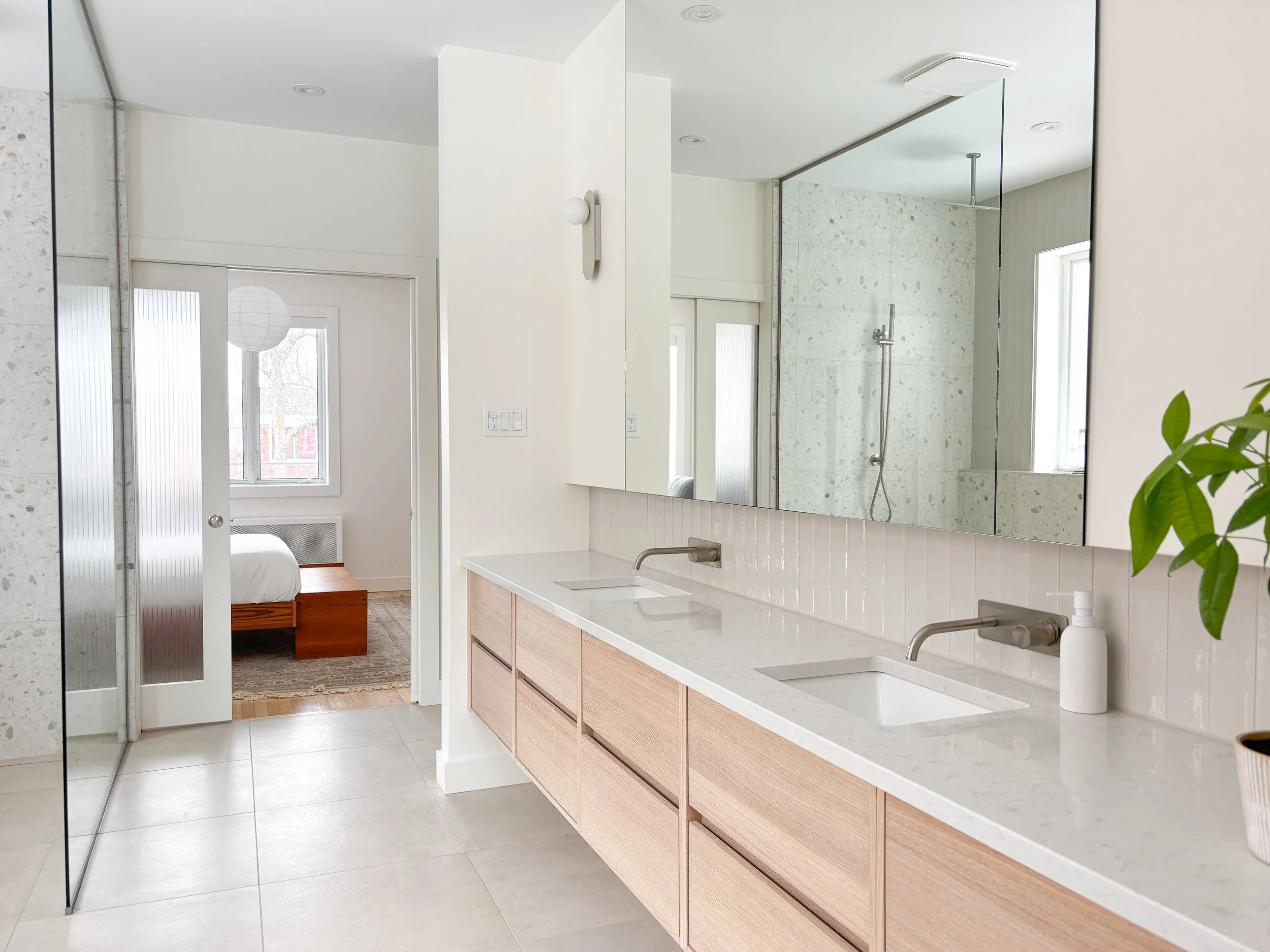Salle de bain moderne avec double lavabo, grand miroir, et vue sur une douche à l'italienne en arrière-plan, chambre visible à travers la porte ouverte.