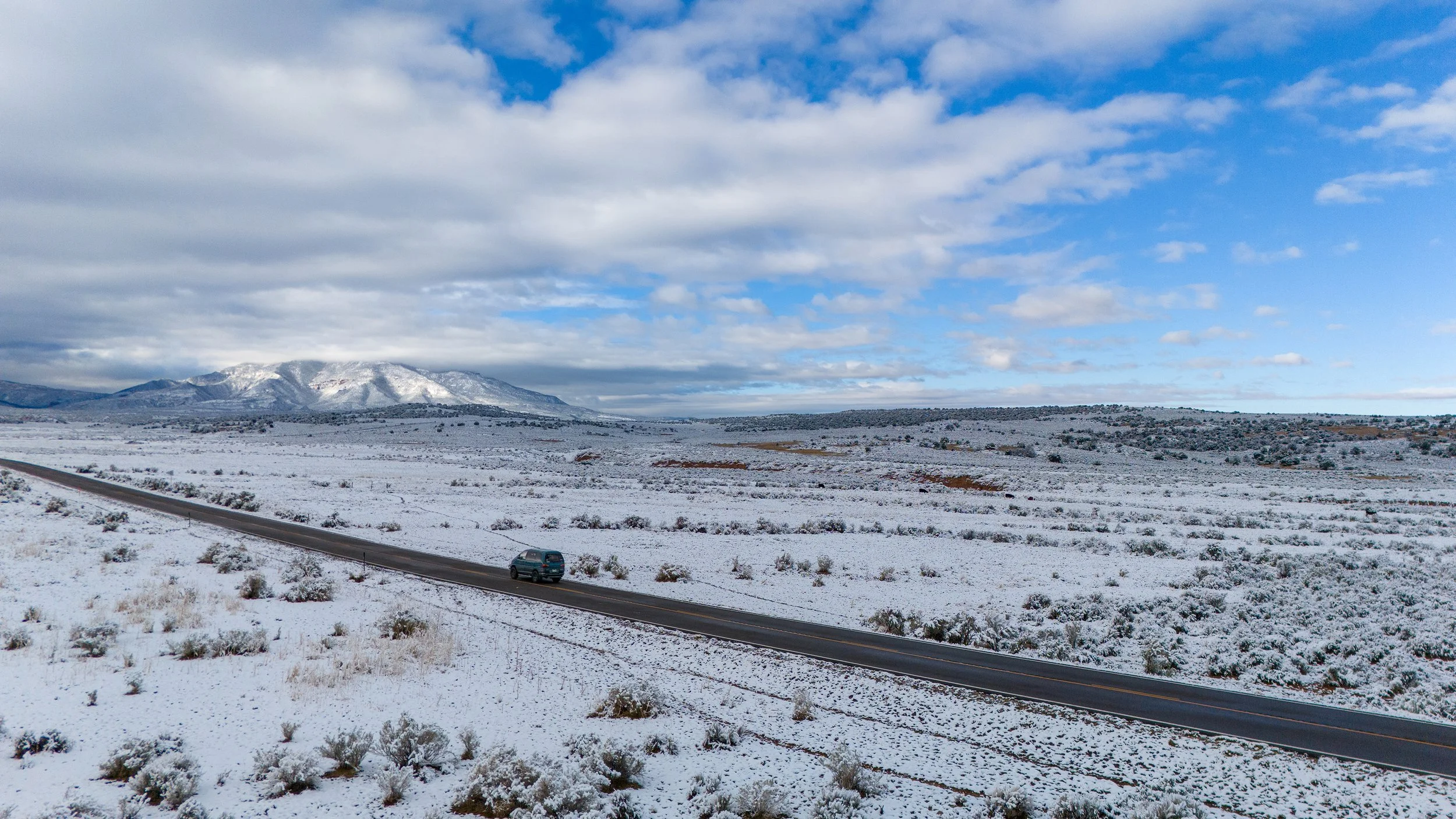 Route de campagne bordée de neige, un véhicule roulant sous un ciel partiellement nuageux, montagnes enneigées au loin.