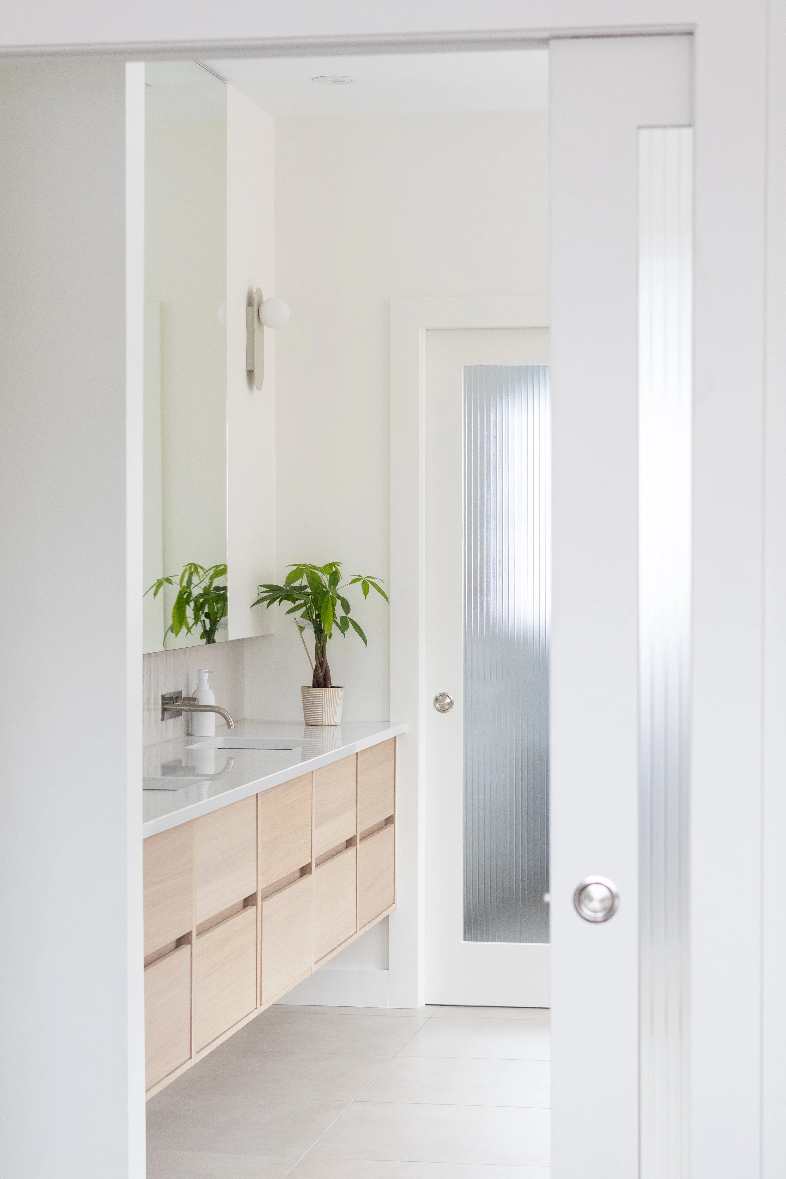 Intérieur d'une salle de bain blanche avec un lavabo, un miroir, un meuble en bois clair, une plante verte en pot, un éclairage mural, et une porte en verre opaque.