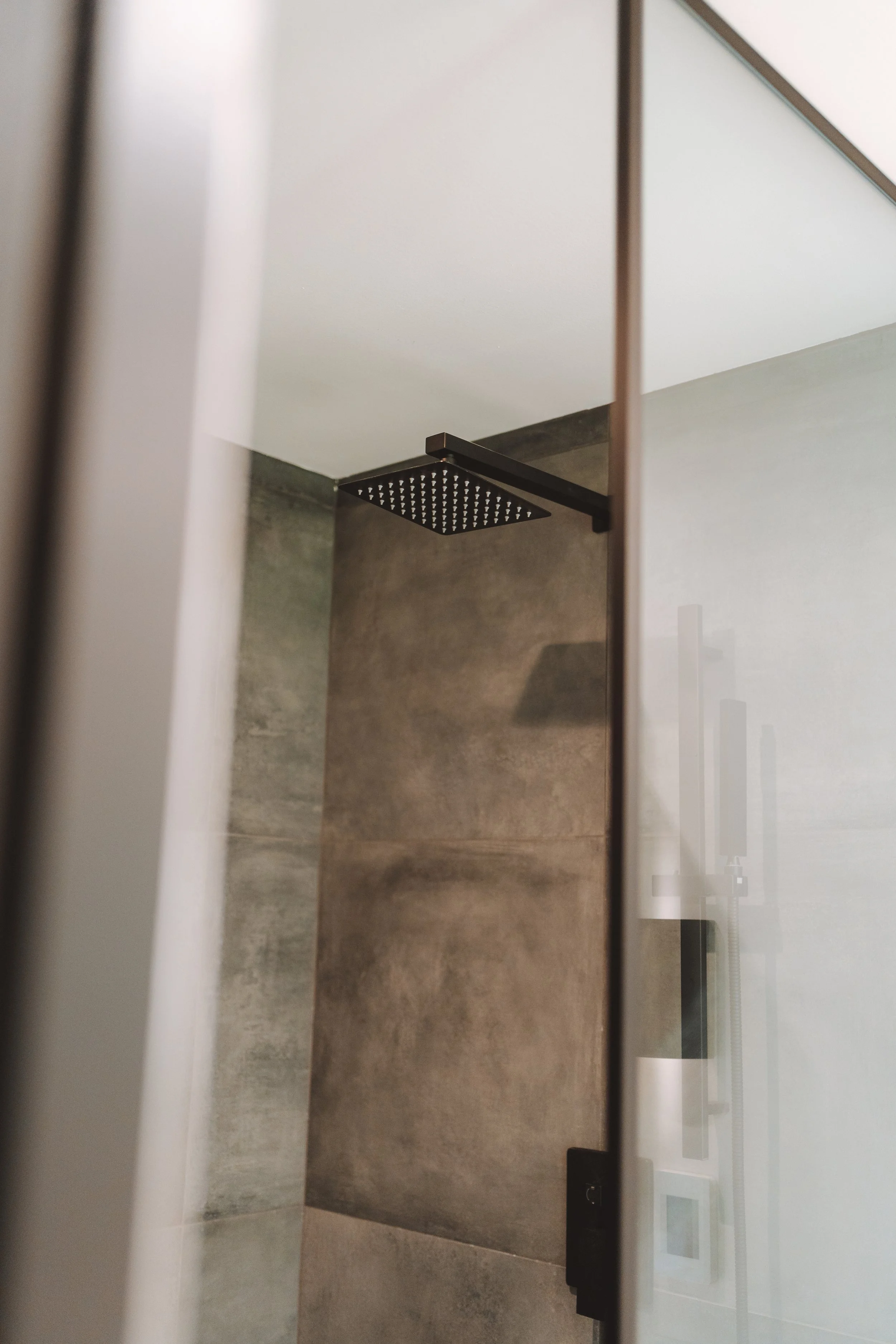 Douche moderne avec un plafond de douche noir, murs en carrelage gris, vue partielle à travers une porte en verre ou une paroi de douche.