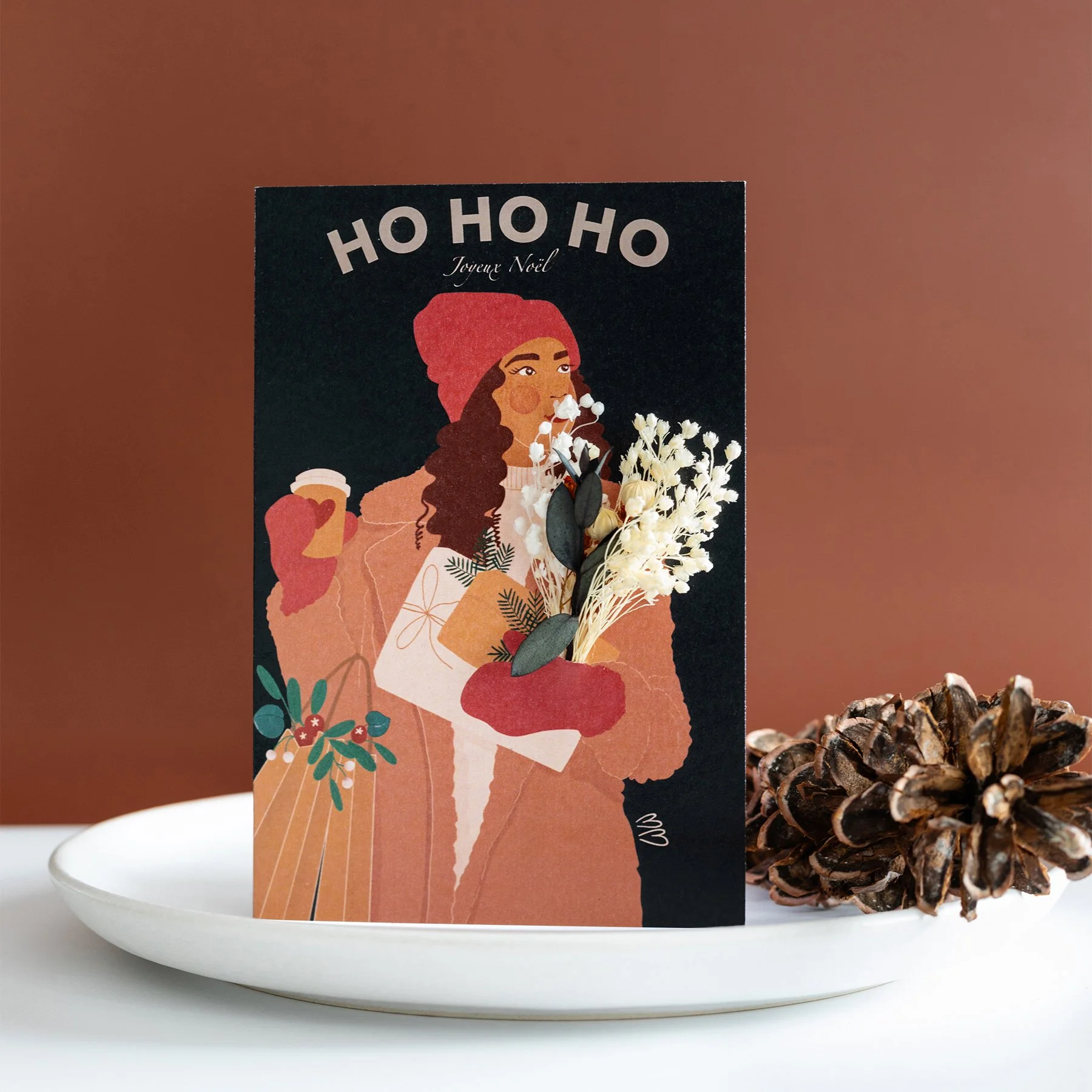 Carte de Noël illustrée avec une femme tenant un bouquet de fleurs blanches, portant un bonnet rouge, avec le texte "Ho Ho Ho Joyeux Noël" en haut, placée sur une assiette blanche avec une pomme de pin à côté.