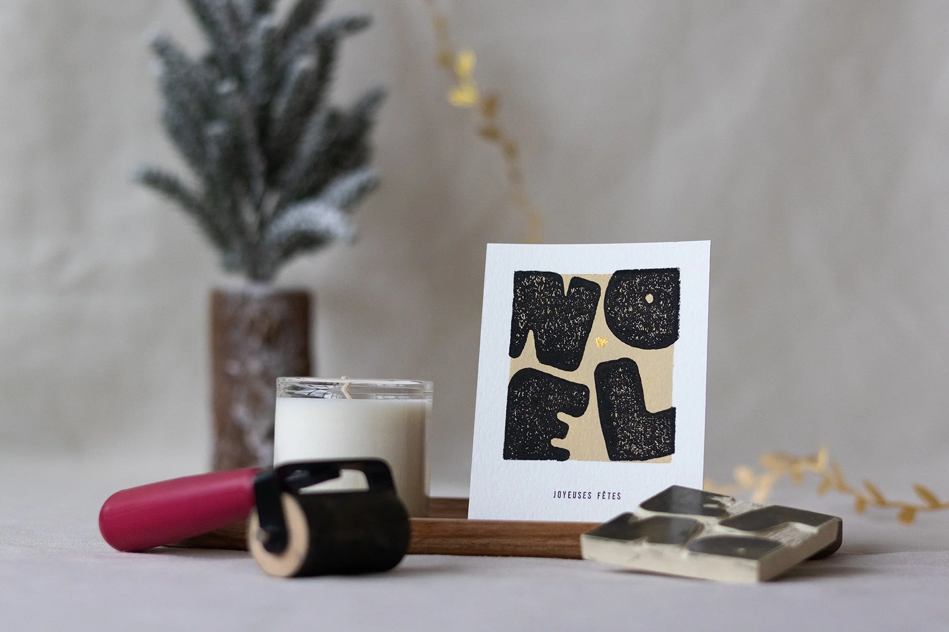 Une carte de vœux avec le mot 'NOEL' en lettres noires, présentée dans un décor de Noël avec un sapin, une bougie et les outils pour la linogravure.