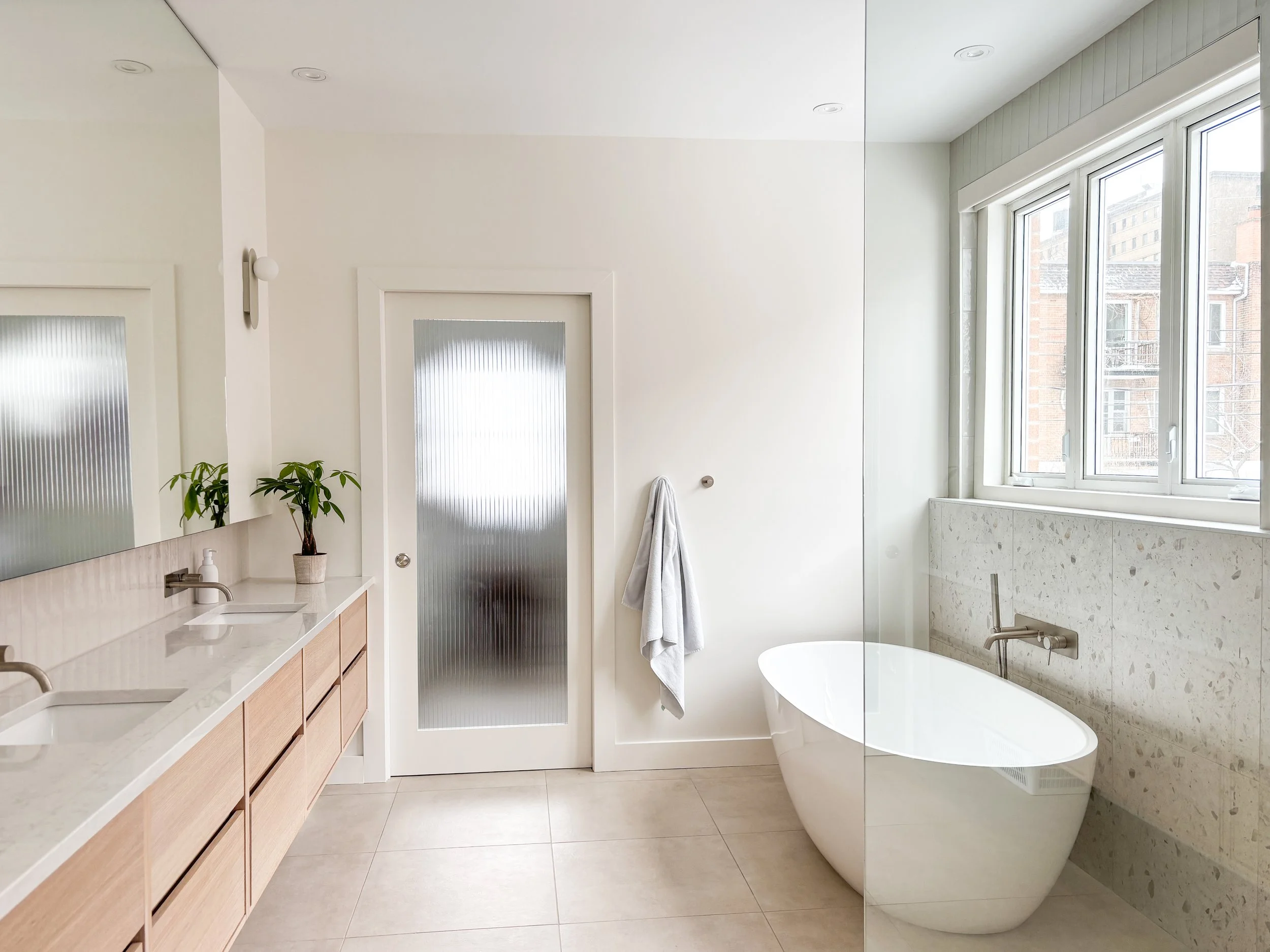 Salle de bain moderne avec double lavabo, miroir, plante, baignoire îlot, grande fenêtre laissant entrer la lumière naturelle, porte en verre givré