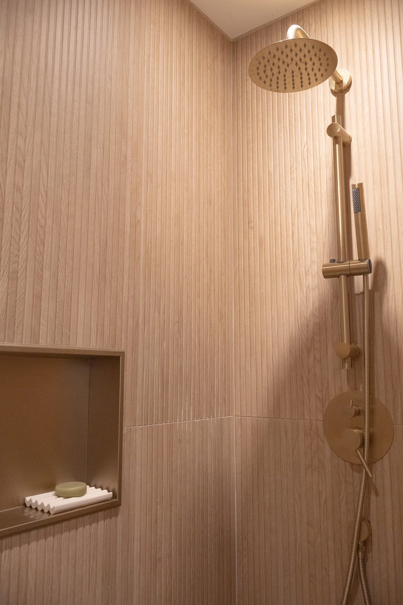 Douche avec pomme de douche dorée ronde, carrelage effet bois avec stries verticales et niche intégrée.