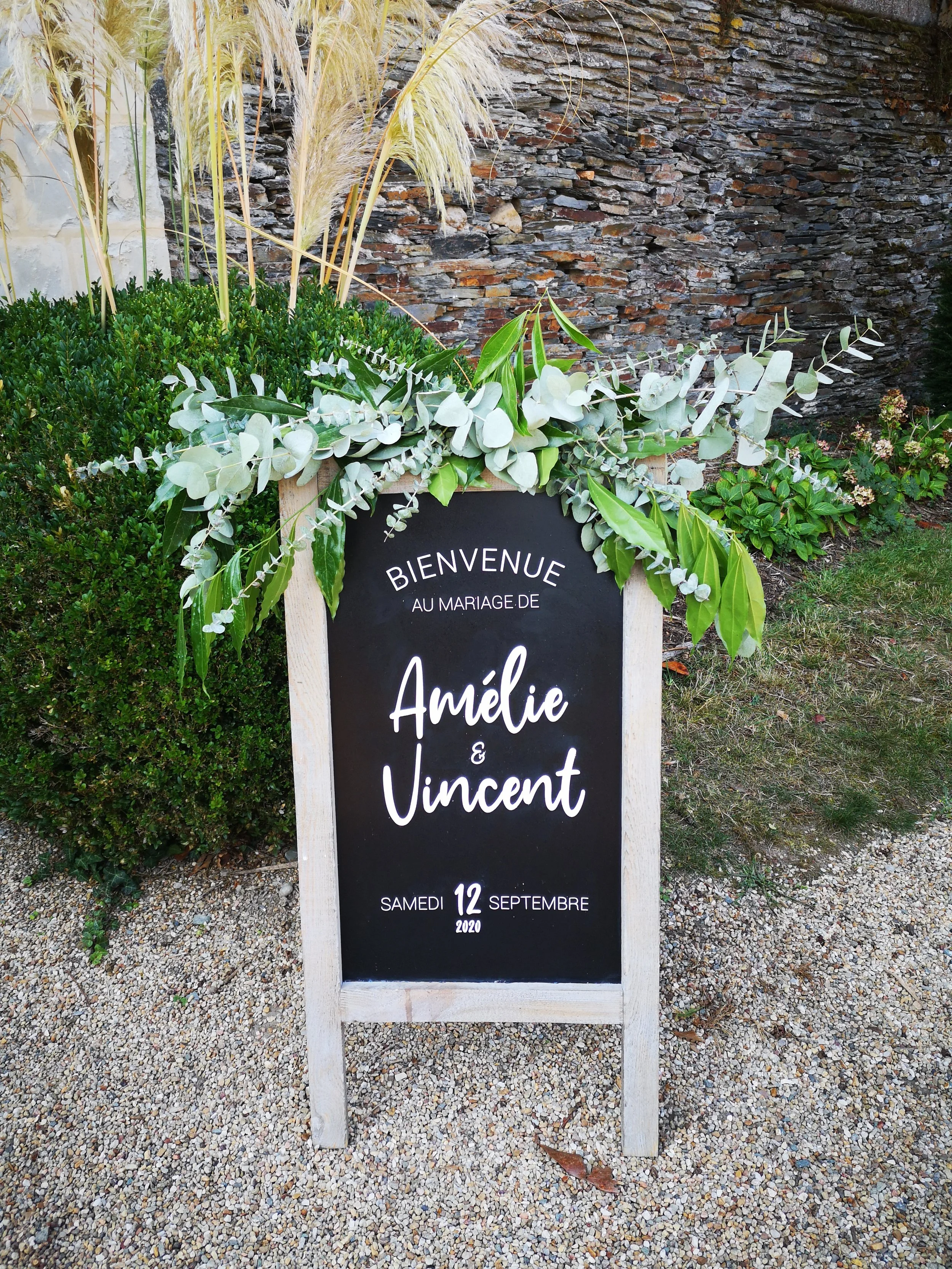 Chalkboard indiquant le mariage et le logo "All is all", décoré de feuillages et placé dans un jardin avec un mur en pierre en arrière-plan.