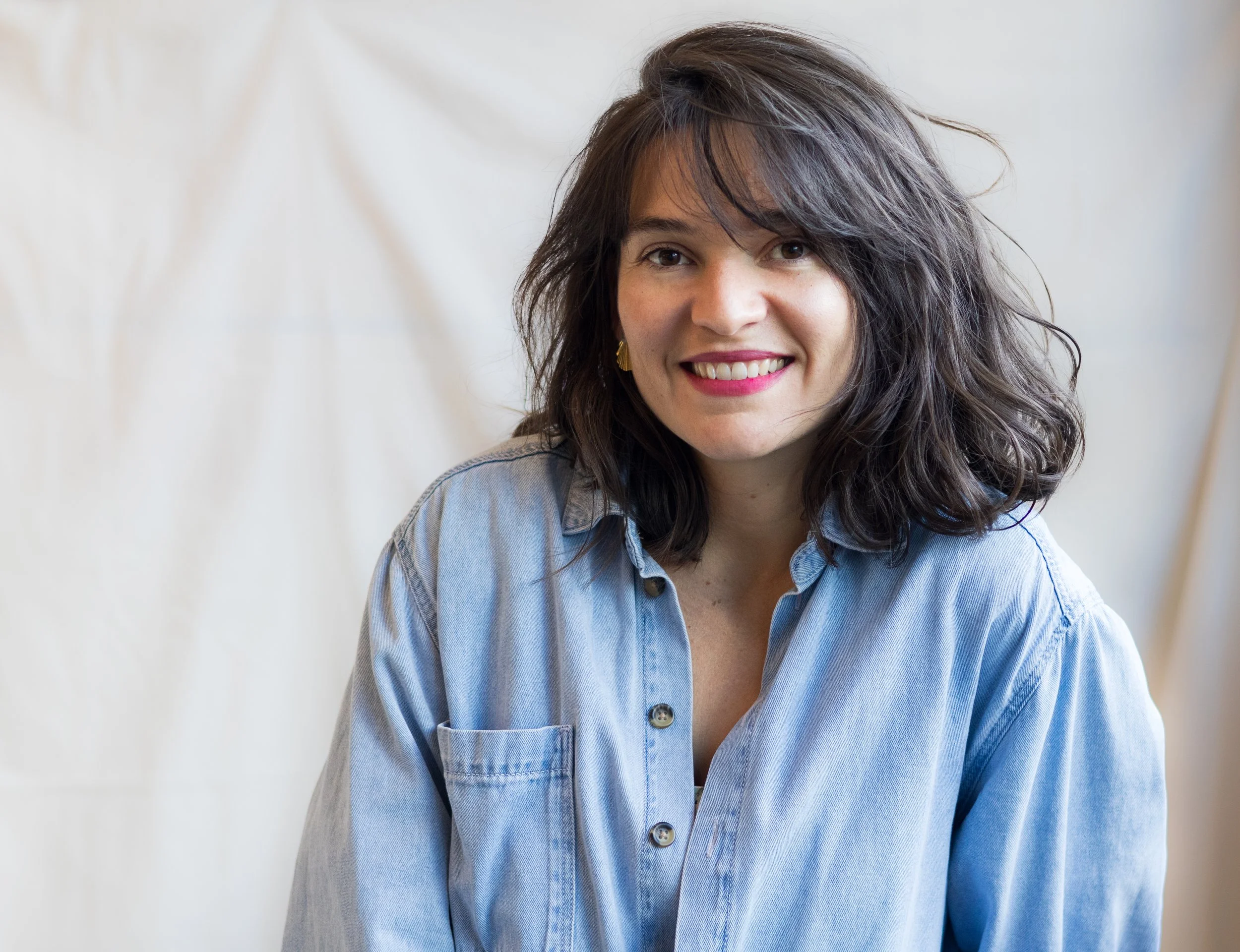 Portrait d'une femme souriante avec des cheveux bruns ondulés, portant une chemise en denim clair.