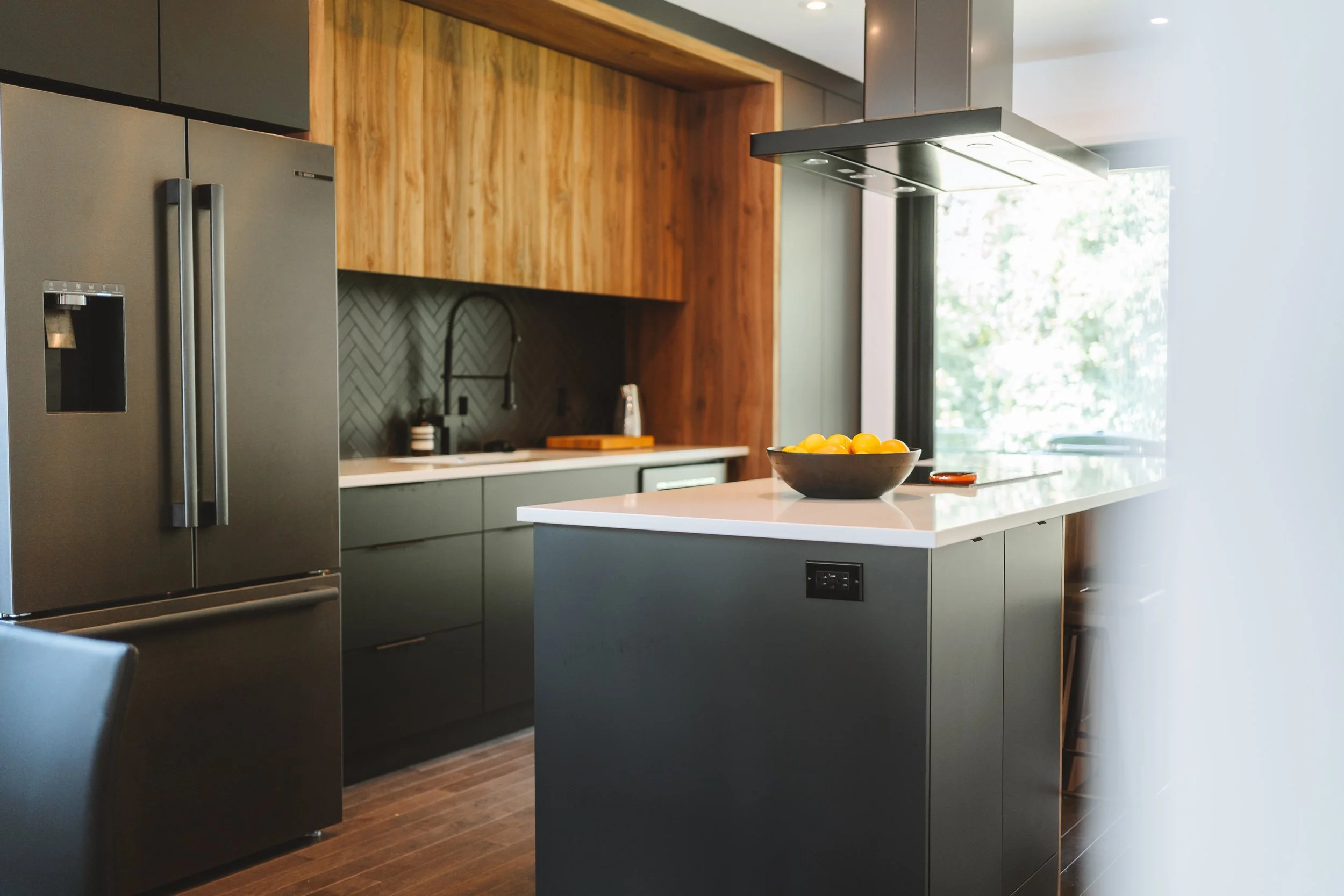 Cuisine moderne avec un îlot central, réfrigérateur double, armoires en bois, plan de travail blanc, arrière-plan avec fenêtres laissant entrer la lumière.