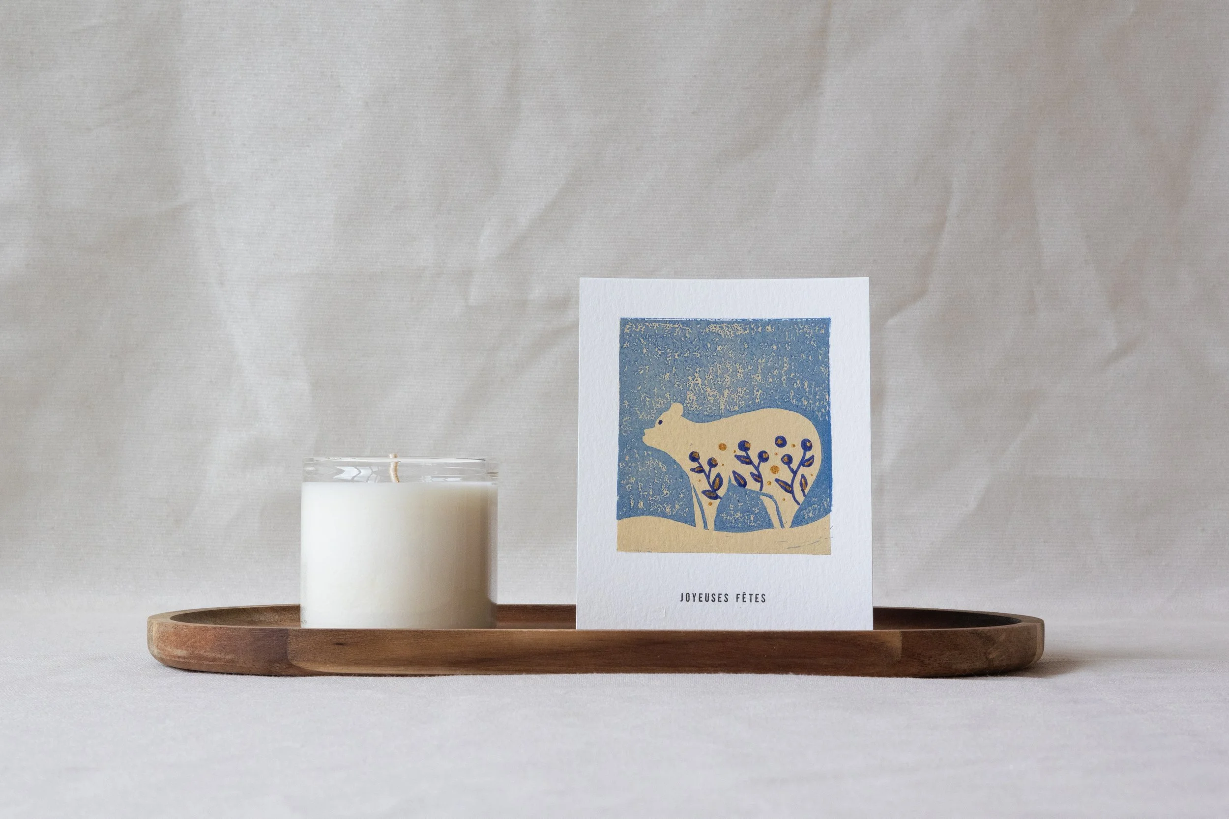 Carte de vœux avec un dessin d'un ours polaire imprimé avec la technique de la linogravure. La carte est posée au côté d'une bougie sur un plateau en bois avec un arrière plan beige.