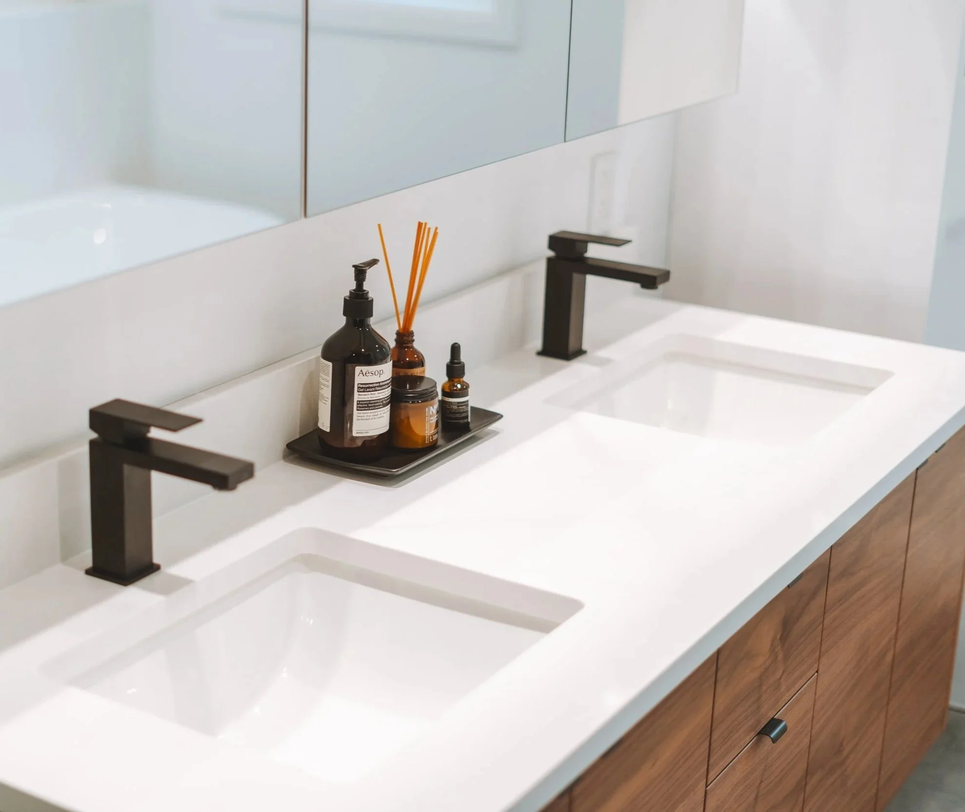 Double vasque en céramique blanche avec un plan de travail en quartz blanc, deux robinets noirs encastrés, une collection de produits de soins et diffuseurs à lierre en haut du meuble en bois naturel.