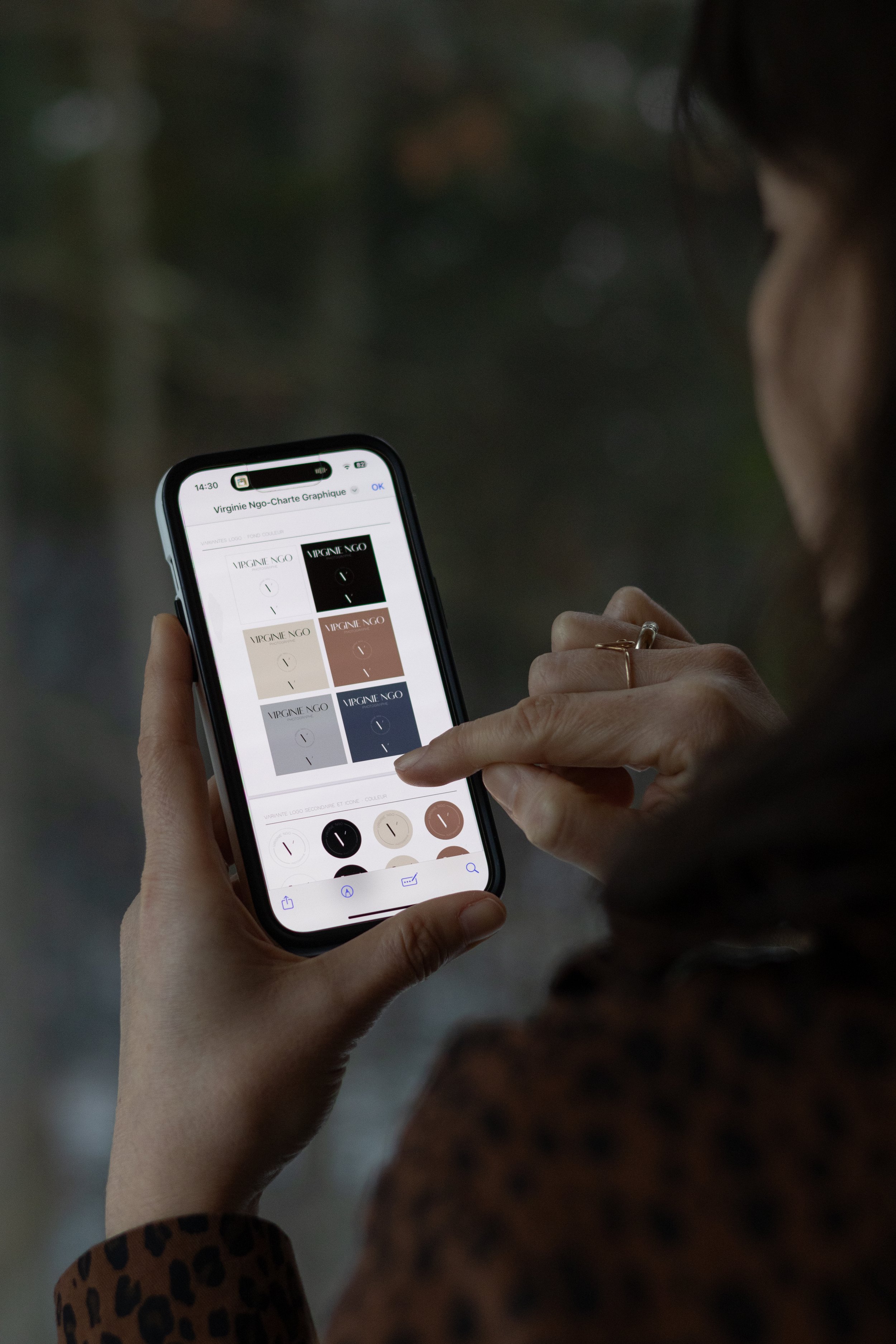 Une personne regarde son téléphone portable, affichant un document avec différents designs de logos pour 'Virginie Ngo'.