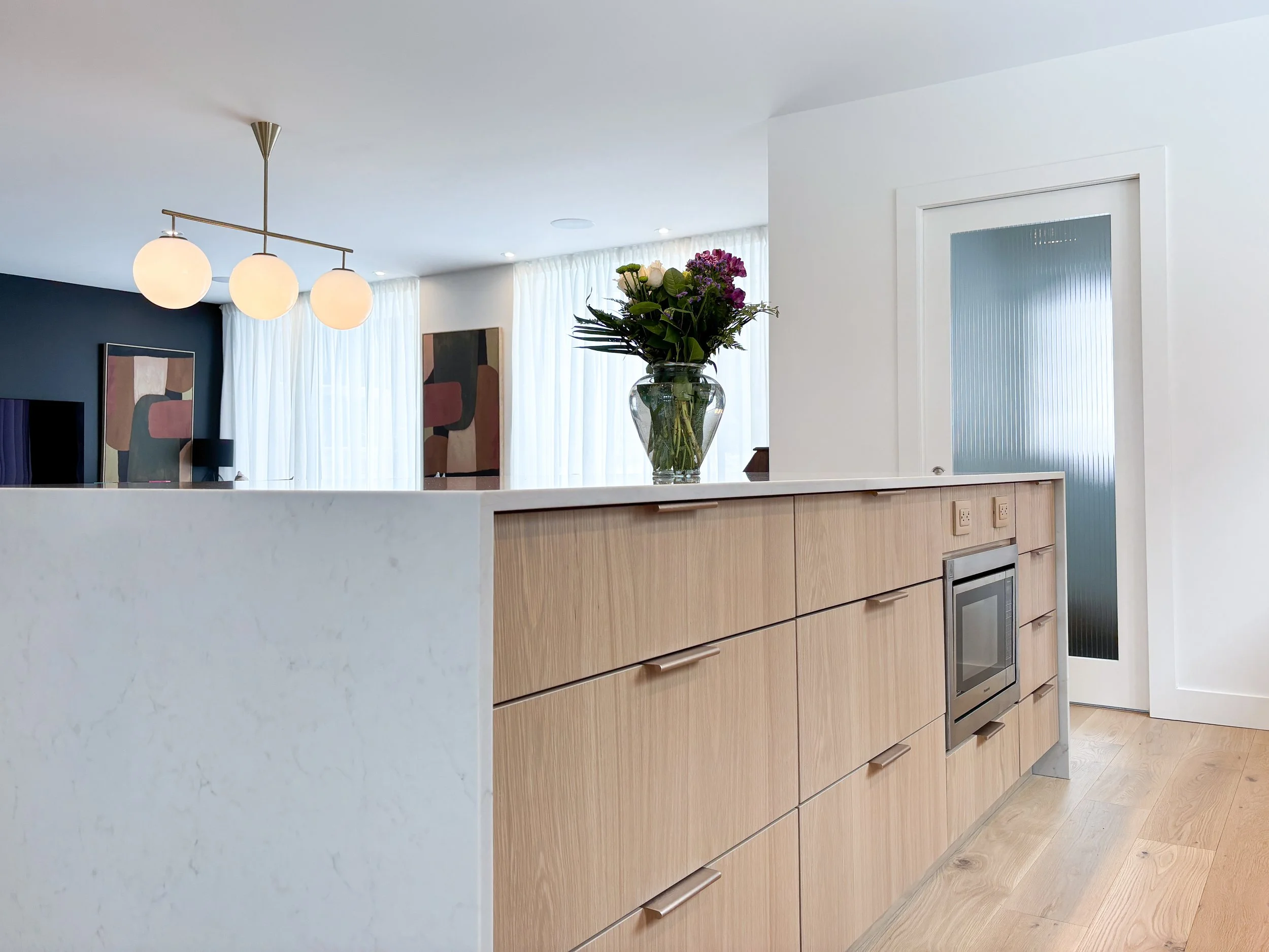 Une cuisine moderne avec un îlot en bois, une vasque de fleurs sur le comptoir, un mur blanc, une porte en verre au fond, et un éclairage suspendu avec trois globes.