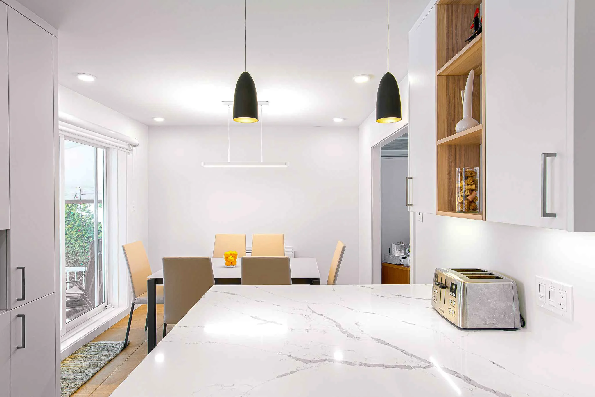 Cuisine moderne avec plan en marbre blanc, planchettes à pain, et lampes suspendues noires. Espace repas avec table et six chaises, porte-fenêtre laissant entrer la lumière.