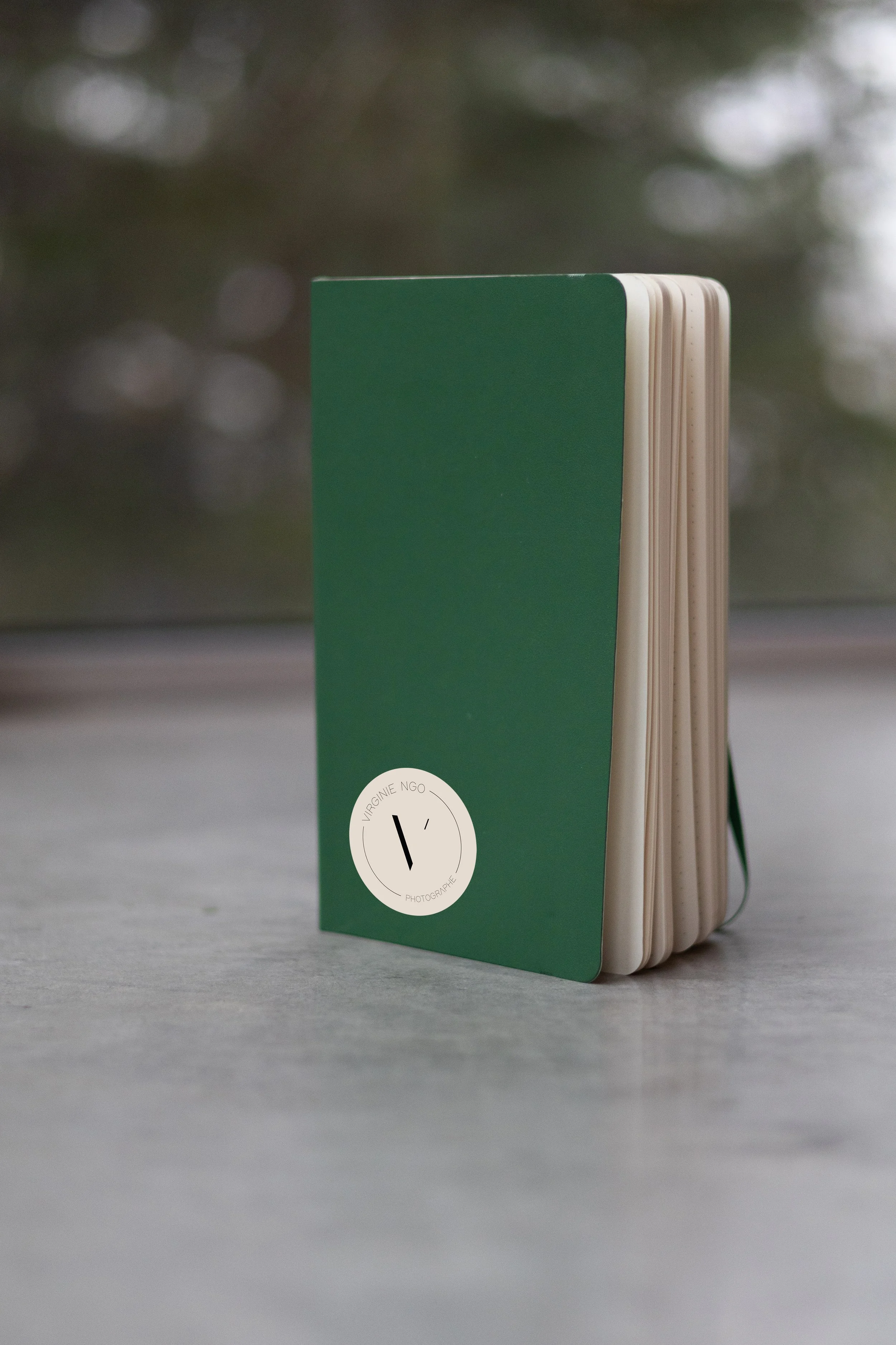 Un carnet vert posé sur une surface de béton avec le sticker de l'iconographie de la photographe Virginie Ngo.